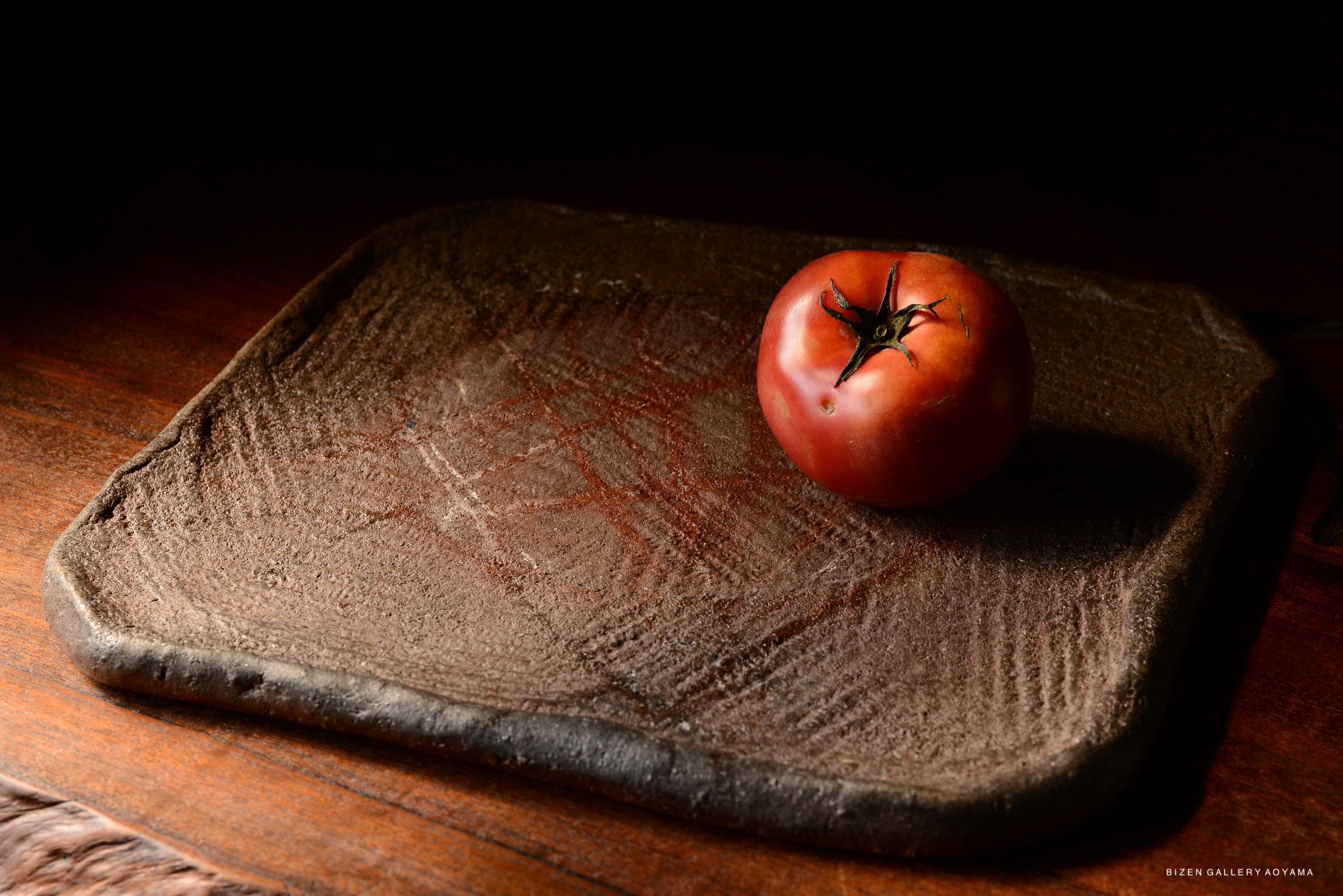 A ripe tomato placed on a textured, dark ceramic plate against a wooden background.