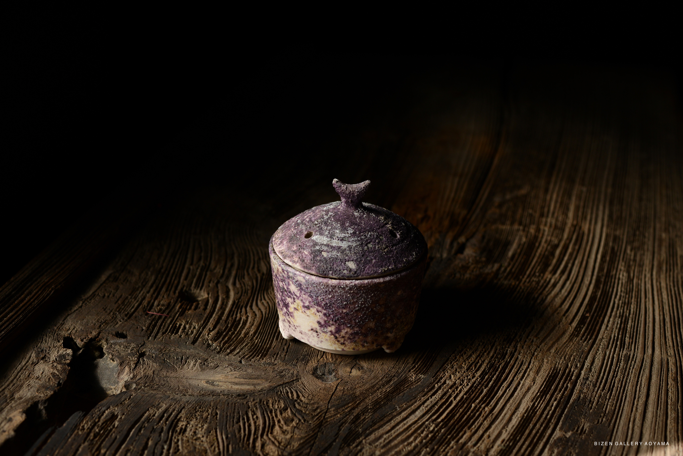 A close-up of a small, purple-tinged ceramic container with a lid, resting on a textured wooden surface in a dimly lit setting.