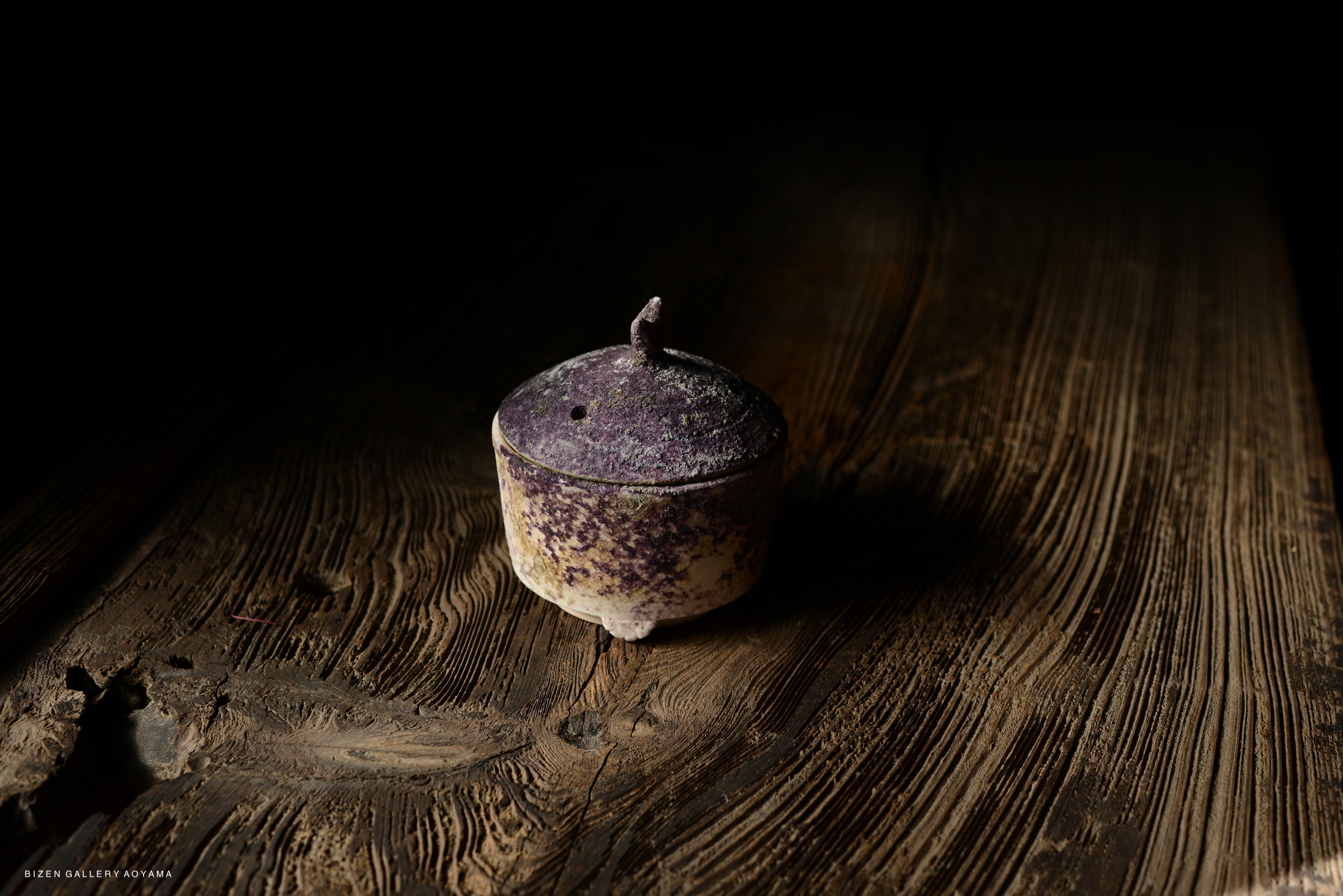 Close-up of a small, purple ash-glazed ceramic vessel on a textured wooden surface, with dark background.