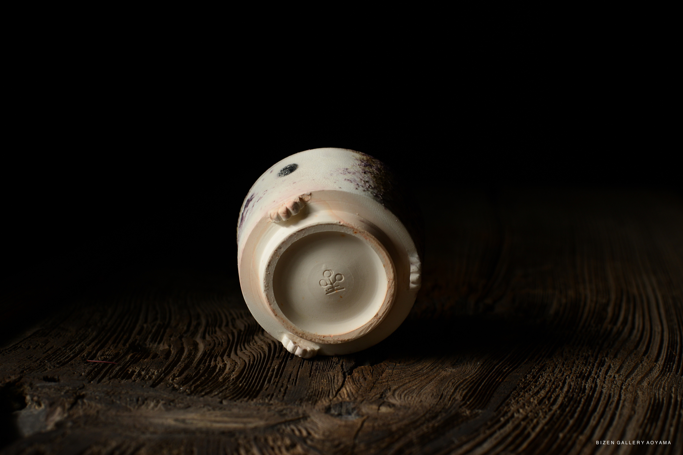Bottom view of a ceramic piece with an ash-glazed surface and small hand-like features, resting on a textured wooden surface.