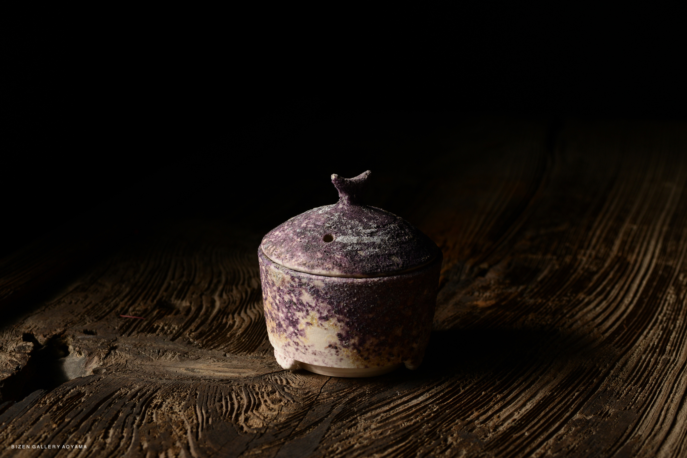 A small purple ceramic container with a lid, featuring a textured surface and an ornamental handle, resting on a wooden table.
