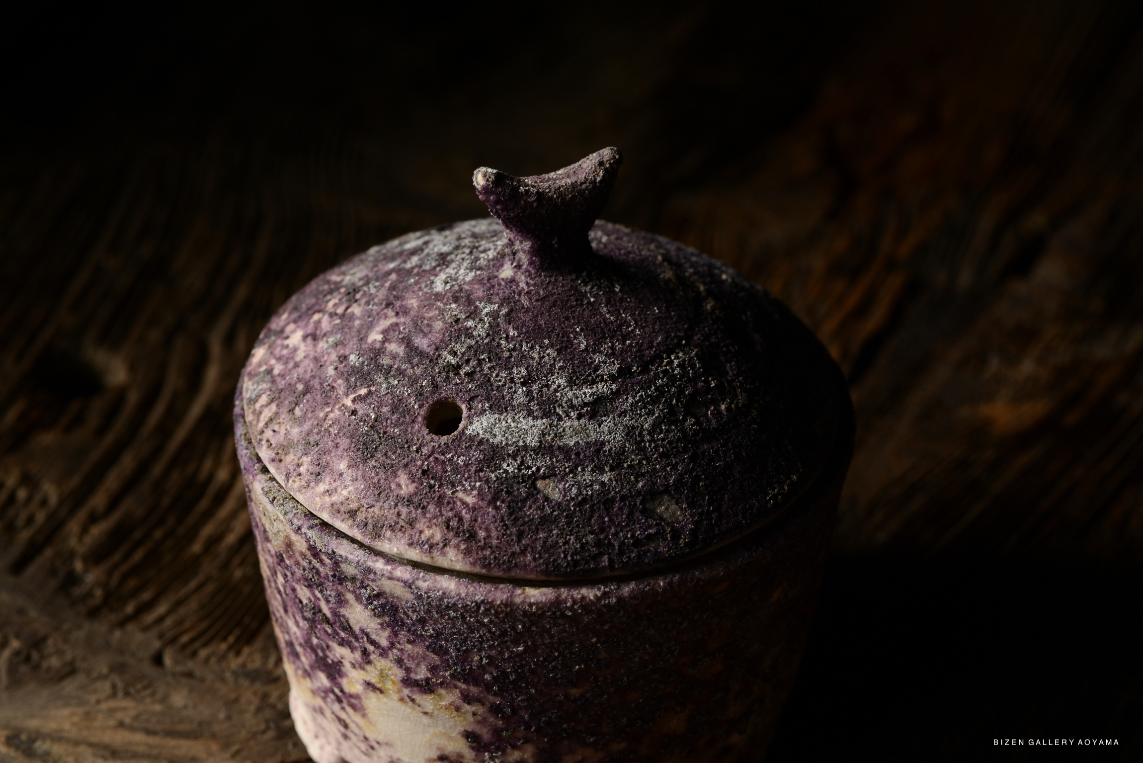 Close-up of a purple-hued ceramic container with a lid, featuring a textured surface and a small protrusion on top.