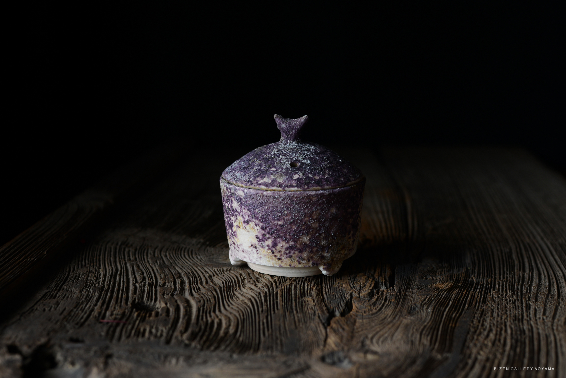 A close-up image of a small, round pottery container with a lid, featuring a textured purple and white surface, placed on a dark wooden table.