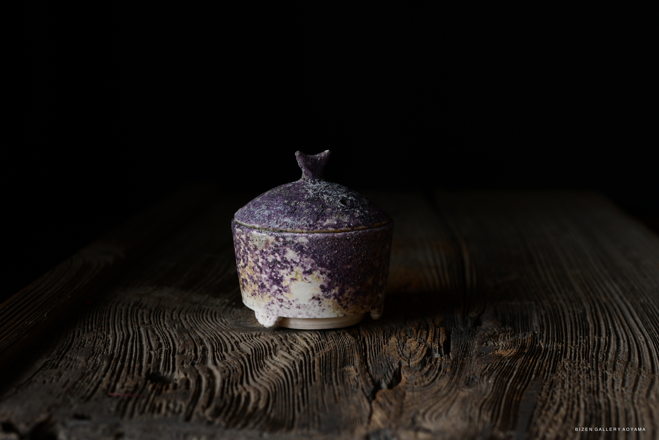 A small ceramic container with a purple speckled lid, placed on a textured wooden surface against a dark background.