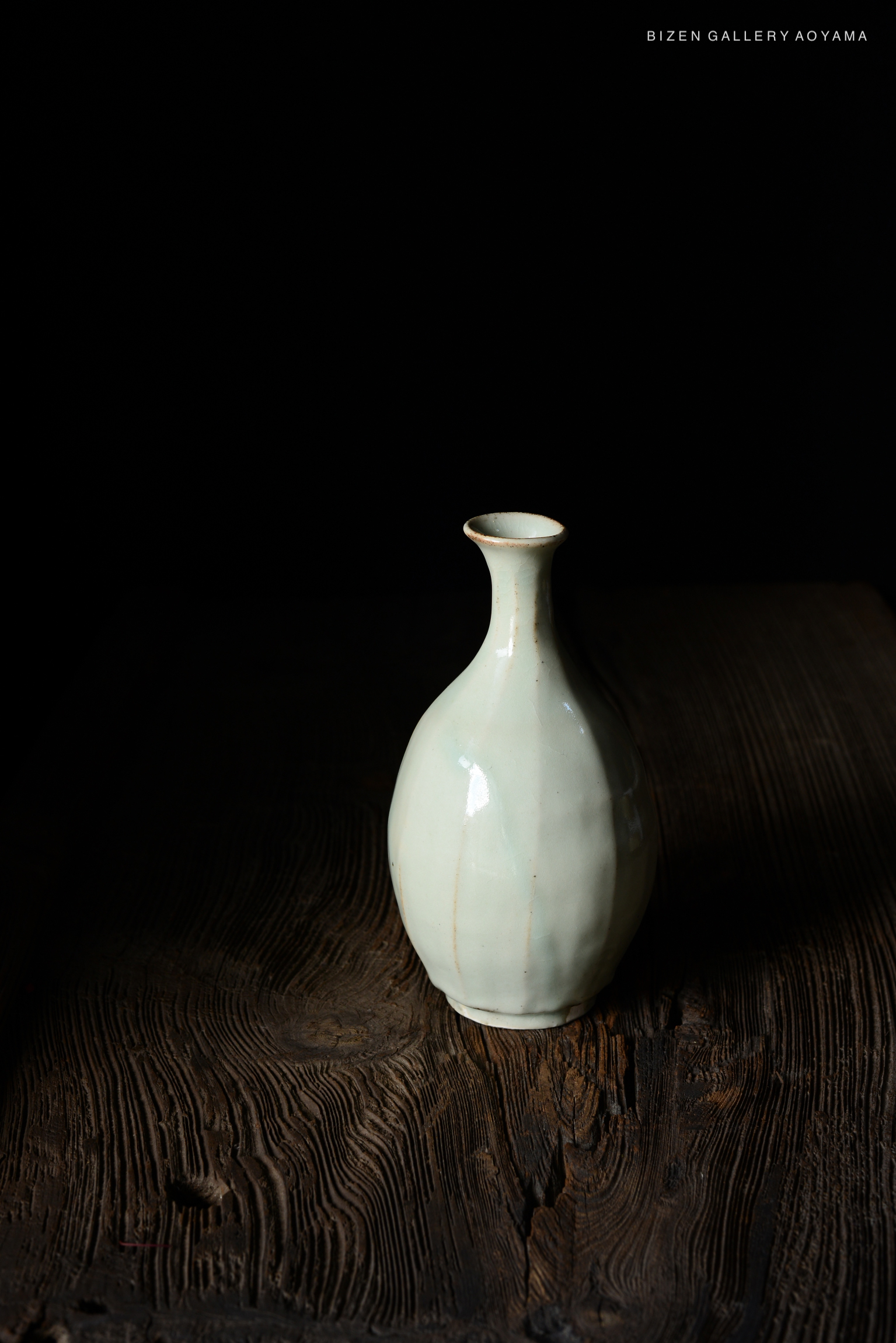 A ceramic vase with a smooth, light green surface and an elongated shape, placed on a wooden table against a dark background.