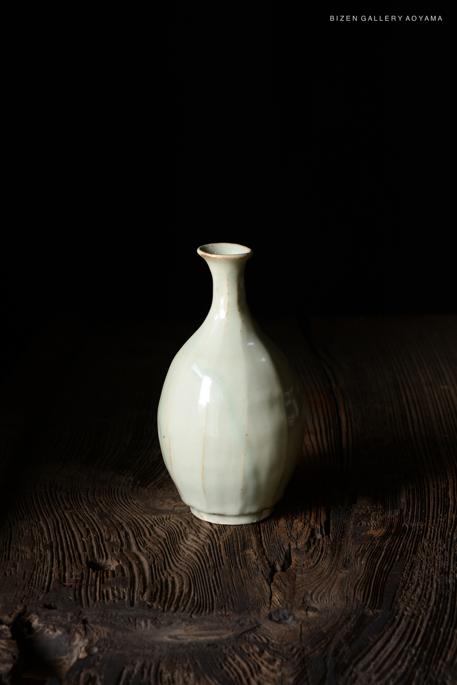 A pale green vase with a narrow neck and a rounded body, resting on a textured wooden surface against a black background.