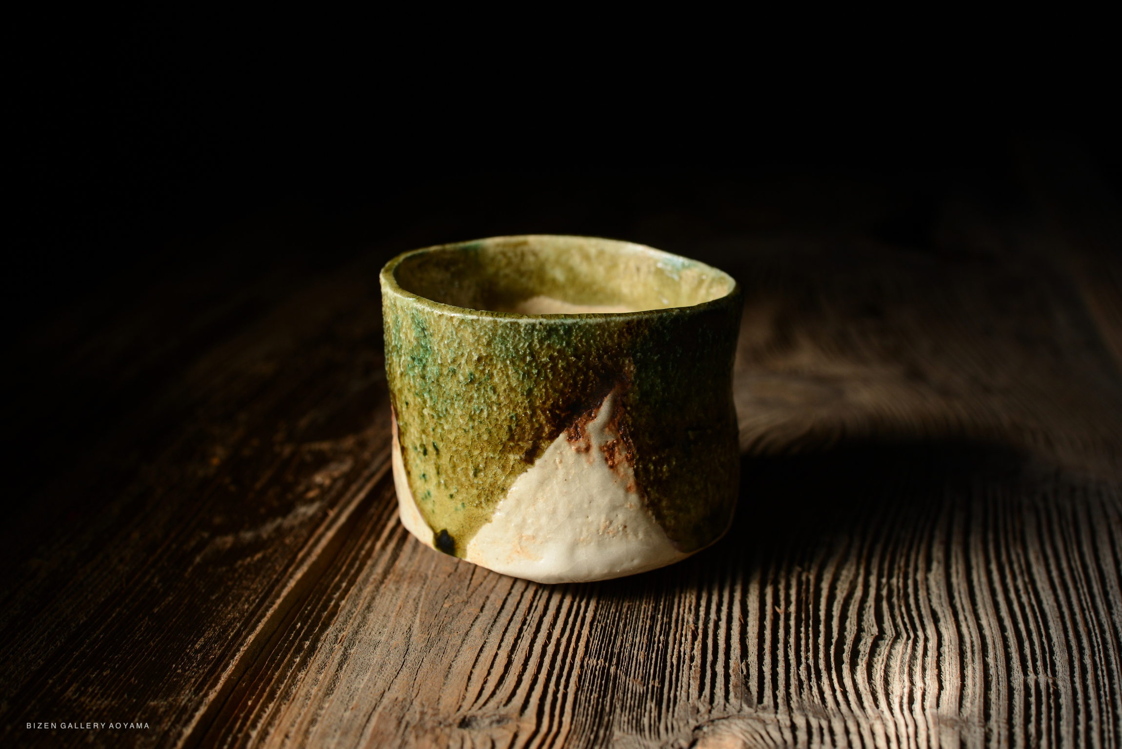 A handcrafted ceramic cup with a textured green and beige surface, positioned on a wooden table with a dark background.