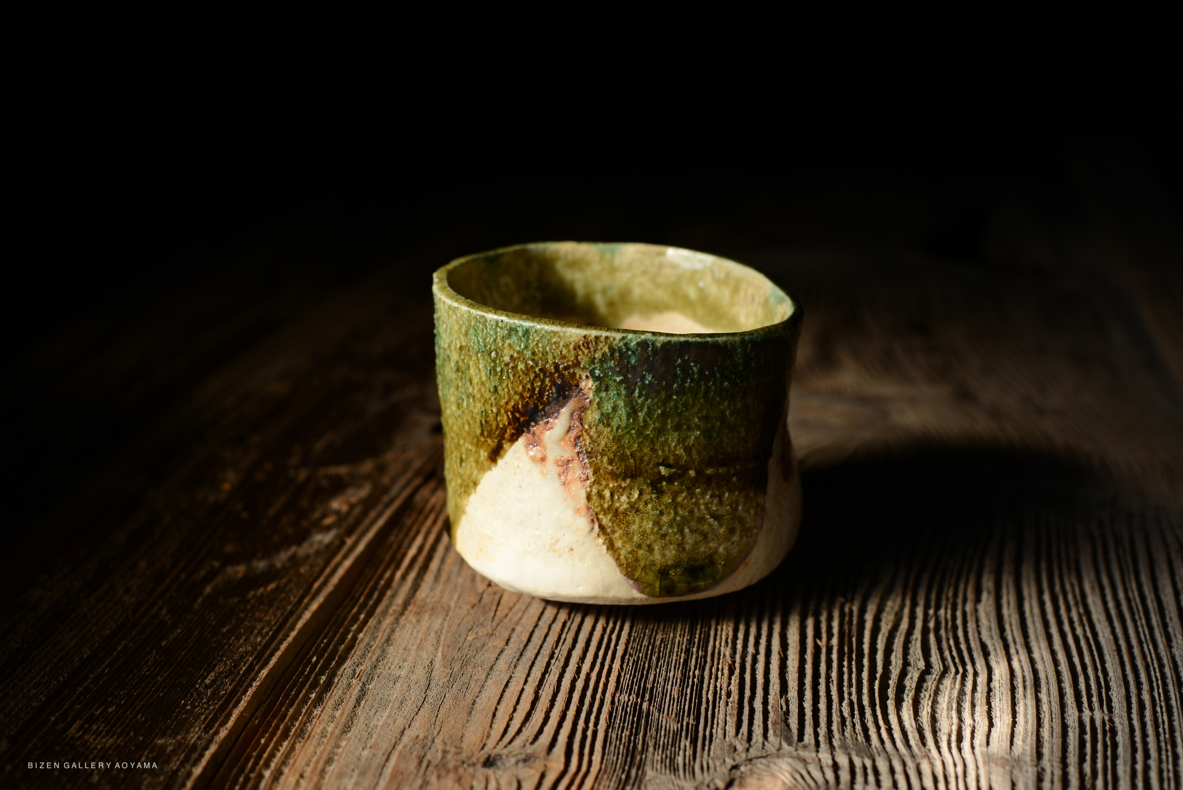 A handcrafted ceramic cup with a textured green and beige surface, sitting on a wooden table.