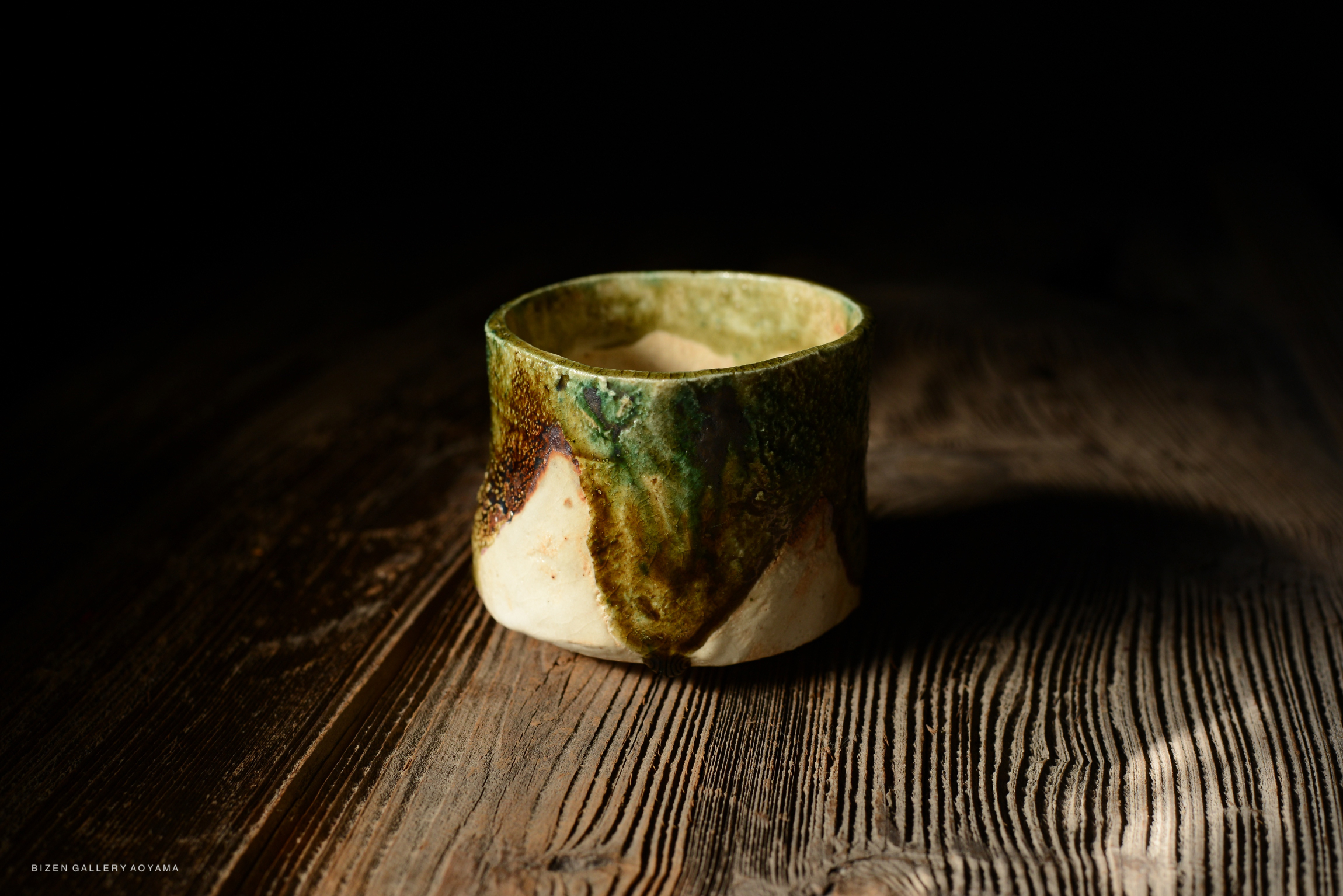 A handcrafted ceramic cup with a green and brown glaze sitting on a textured wooden surface.