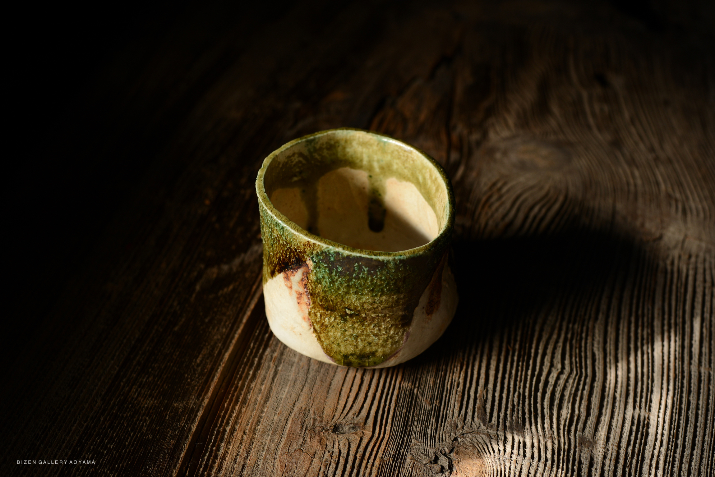 A handcrafted ceramic cup with a textured green and brown glaze, placed on a wooden surface.