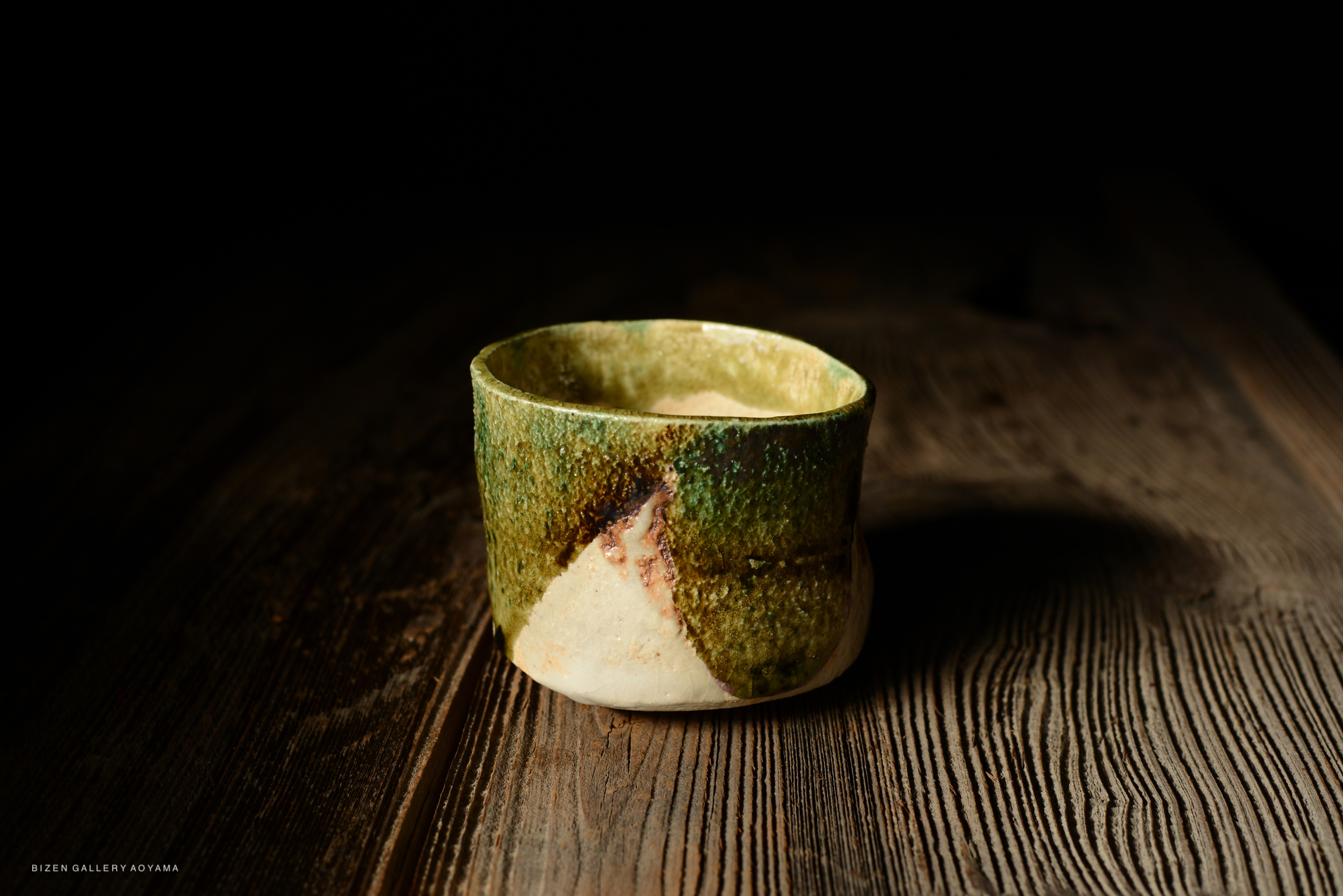 A close-up view of a handcrafted ceramic cup with a green and beige glaze, resting on a textured wooden surface.