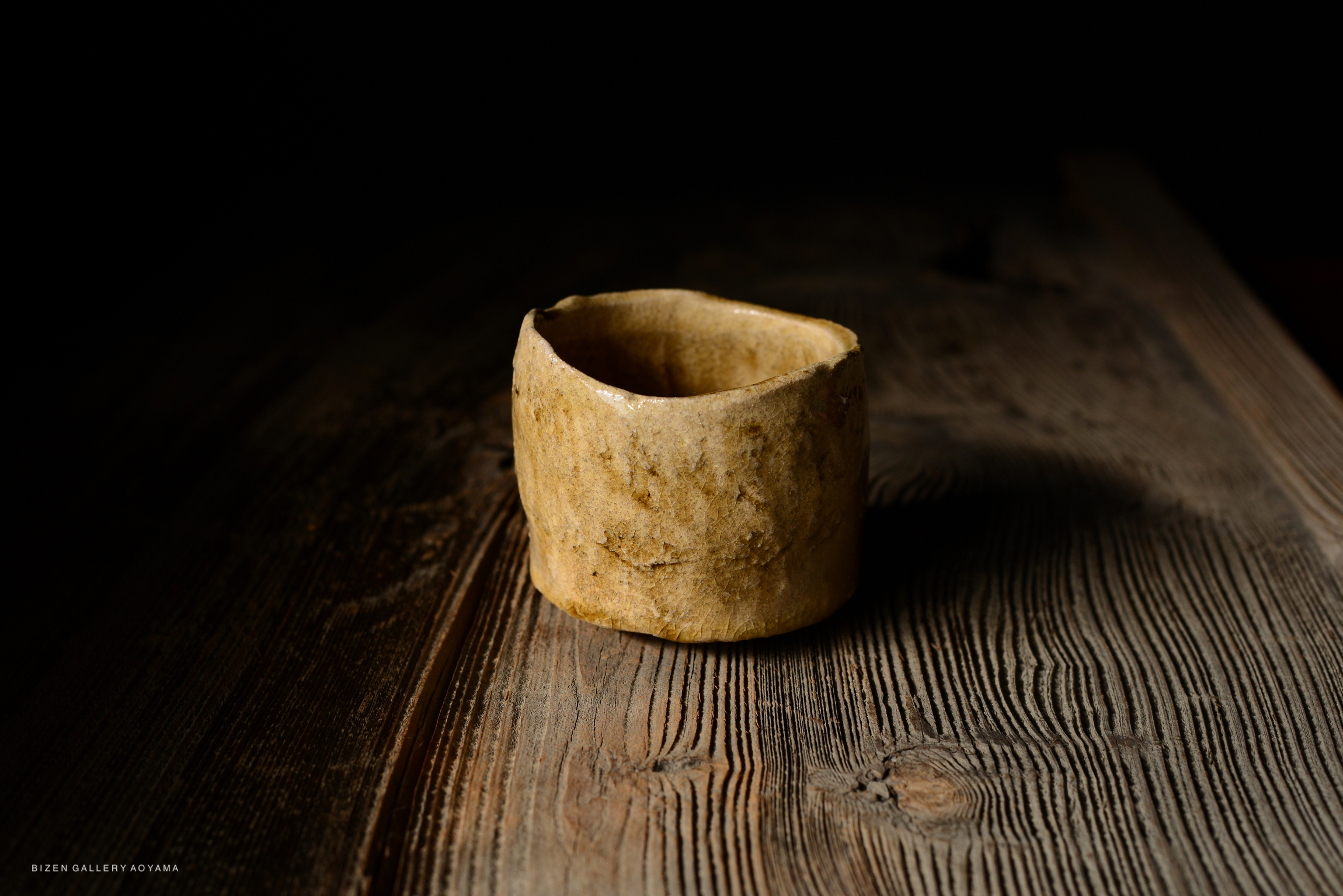A close-up of a round, unglazed ceramic cup resting on a wooden surface, with dark background.