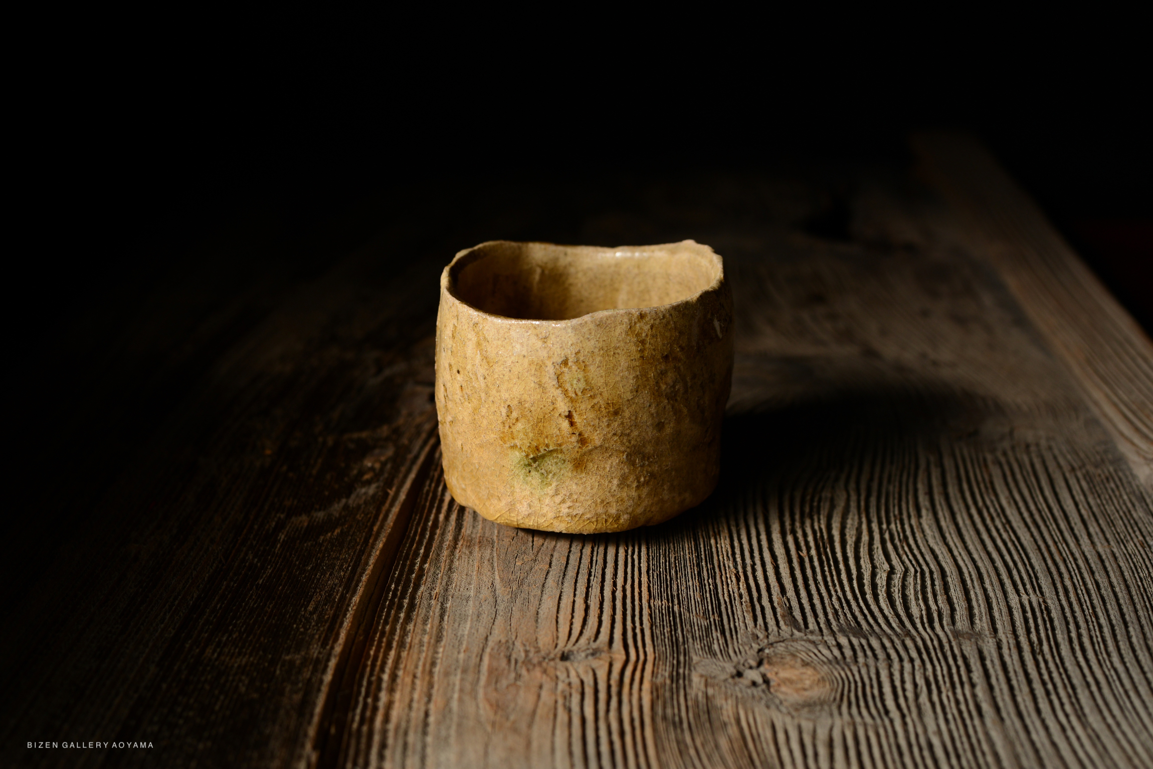 A rustic, handmade ceramic cup with an uneven rim, placed on a wooden table with a dark background.