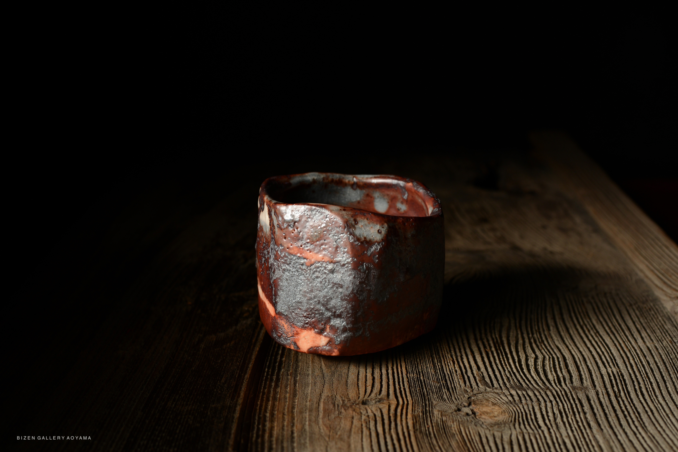 A uniquely shaped ceramic cup with a blend of red and gray tones sits on a wooden surface, illuminated against a dark background.