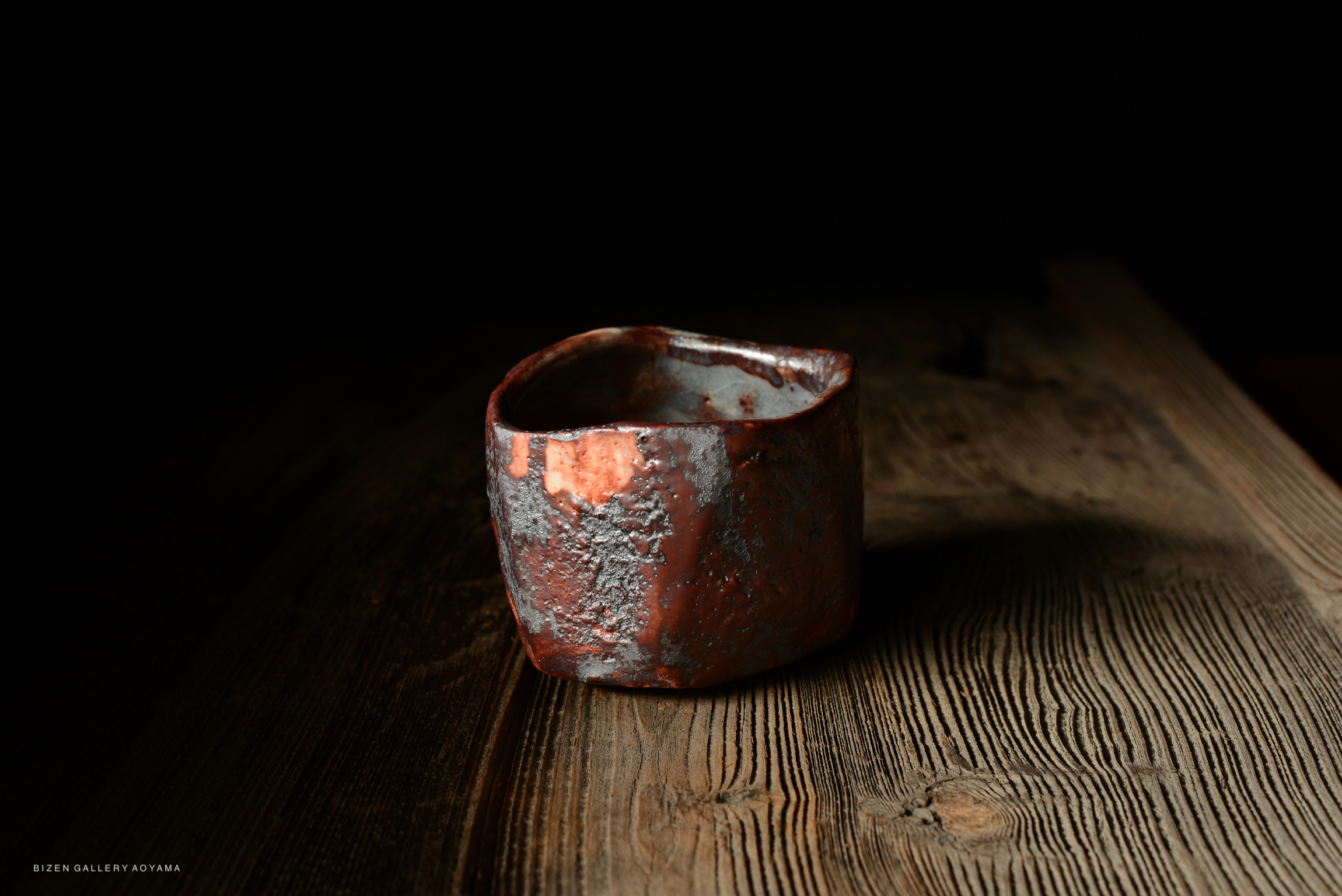 A close-up image of a handmade ceramic cup with an irregular shape, featuring textured surfaces and reddish-brown hues, placed on a rustic wooden table.