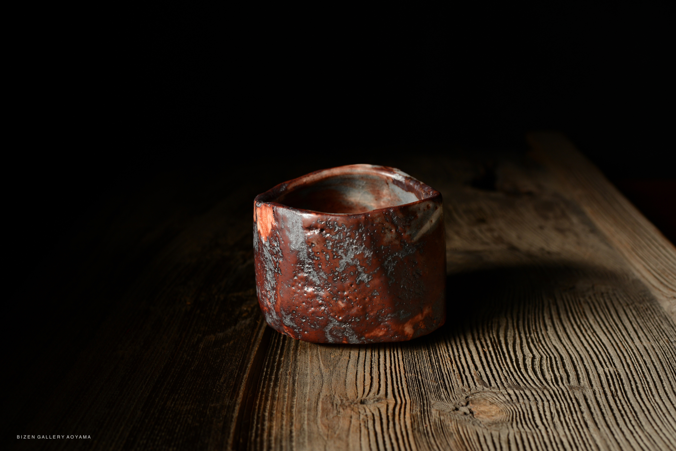 A rustic ceramic bowl with a textured surface in shades of red and gray, placed on a wooden table against a dark background.