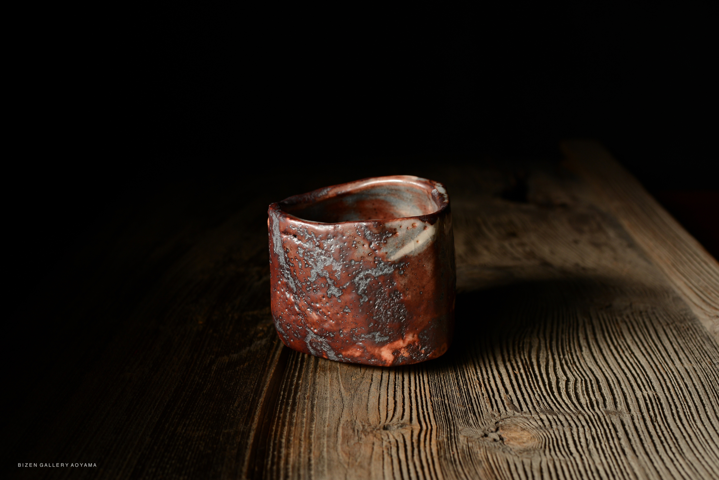 A uniquely shaped ceramic cup with a reddish-brown glaze, featuring textured patterns, resting on a rustic wooden surface under low light.