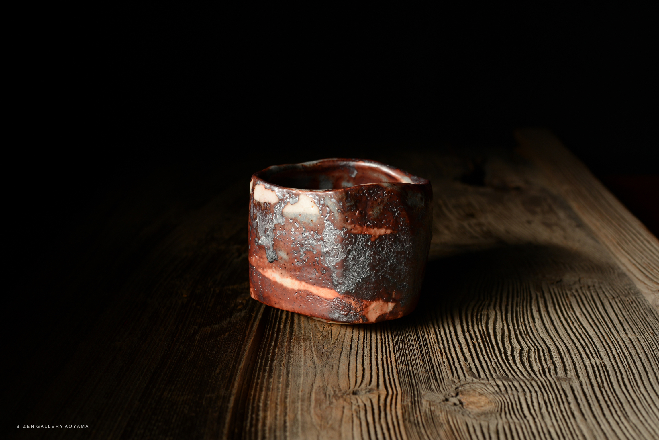 A rustic ceramic cup with a textured surface and varied earthy tones, resting on a wooden table with soft lighting.