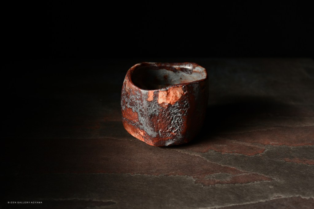 A handcrafted ceramic tea bowl with a textured, reddish-brown surface, set against a dark background.