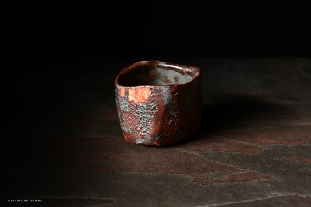 A close-up of a unique, handcrafted ceramic bowl with a textured, reddish-brown exterior and a light-colored interior, set against a dark background.