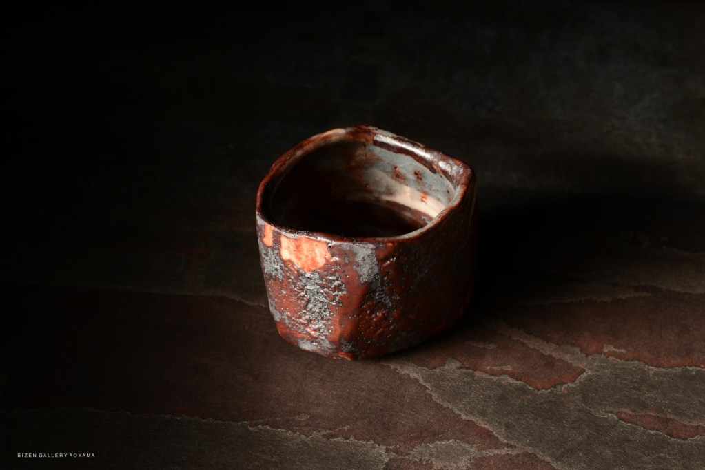 A unique red ceramic vessel with a rough texture and irregular shape, placed on a dark surface.