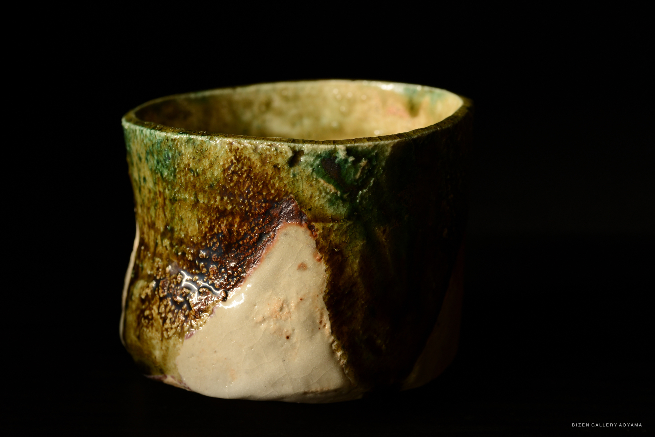 A close-up view of a uniquely shaped ceramic bowl with a textured surface, showcasing a blend of earthy colors including green, brown, and beige against a dark background.