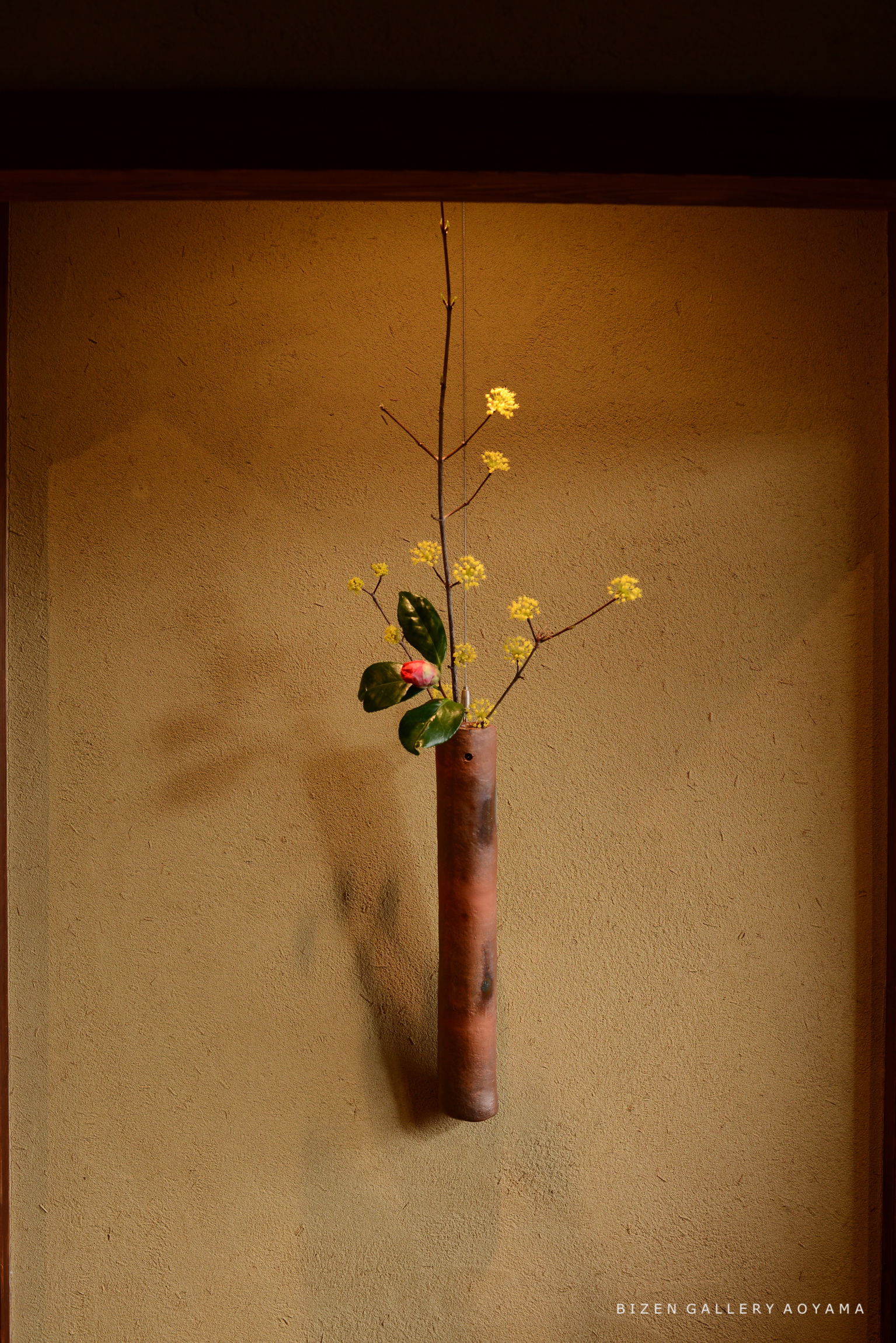 茶色の壁に掛けられた筒状の花瓶に、黄色い花と緑の葉が生けられている様子。