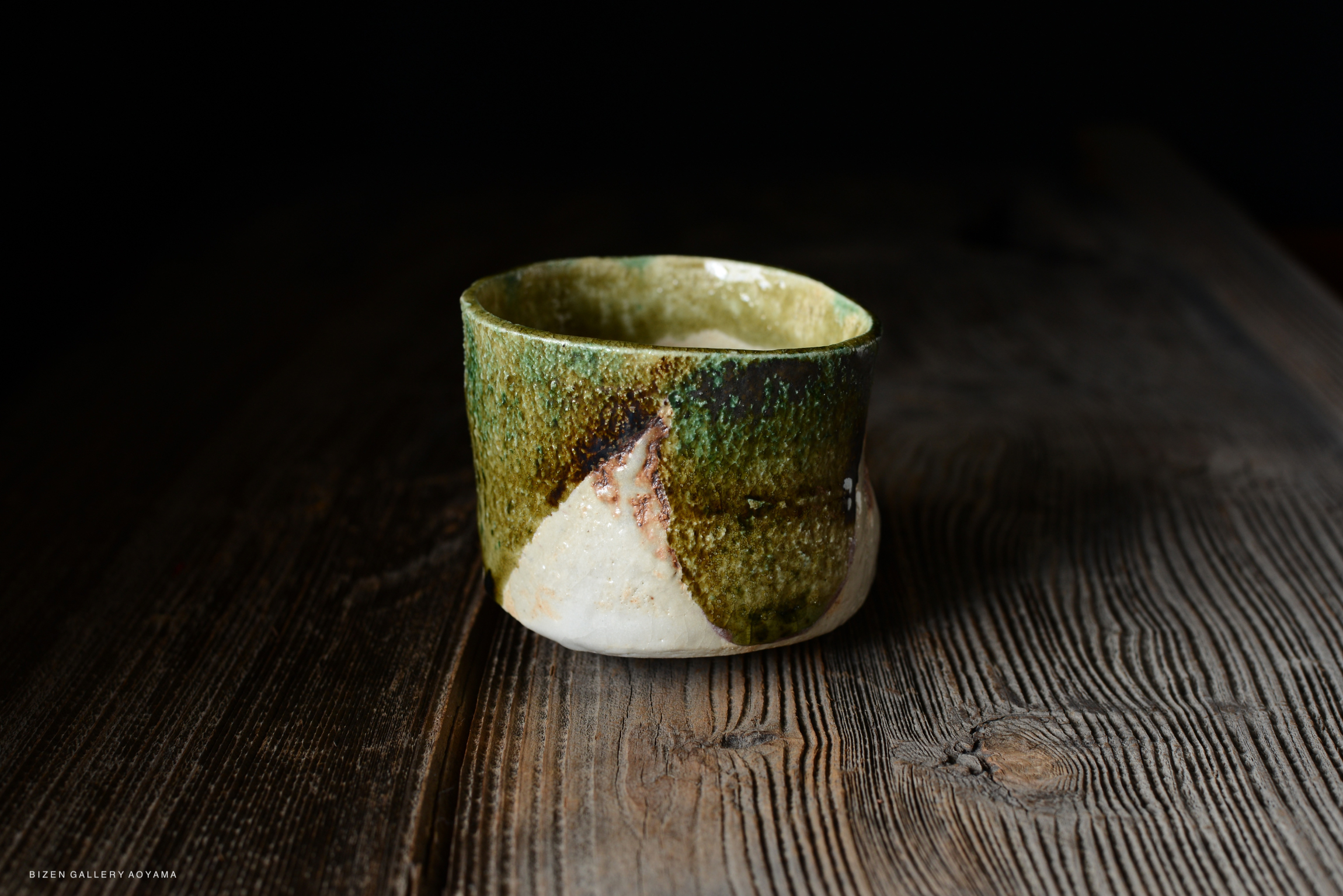 A unique ceramic cup with a green and beige glaze, featuring a textured surface, placed on a wooden table.