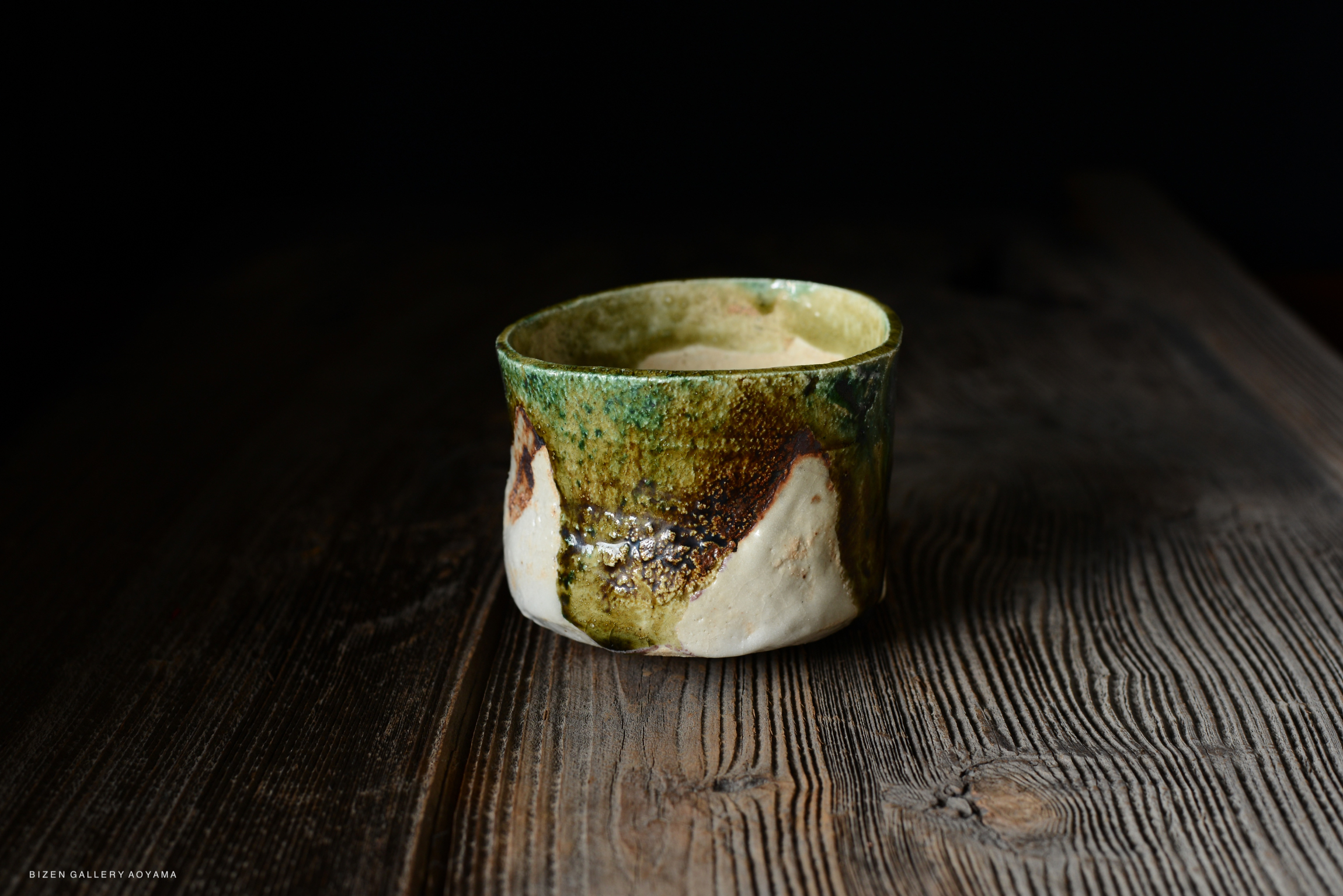 A close-up view of a handmade ceramic bowl with a textured surface, featuring a mix of green and earthy tones, placed on a wooden table.