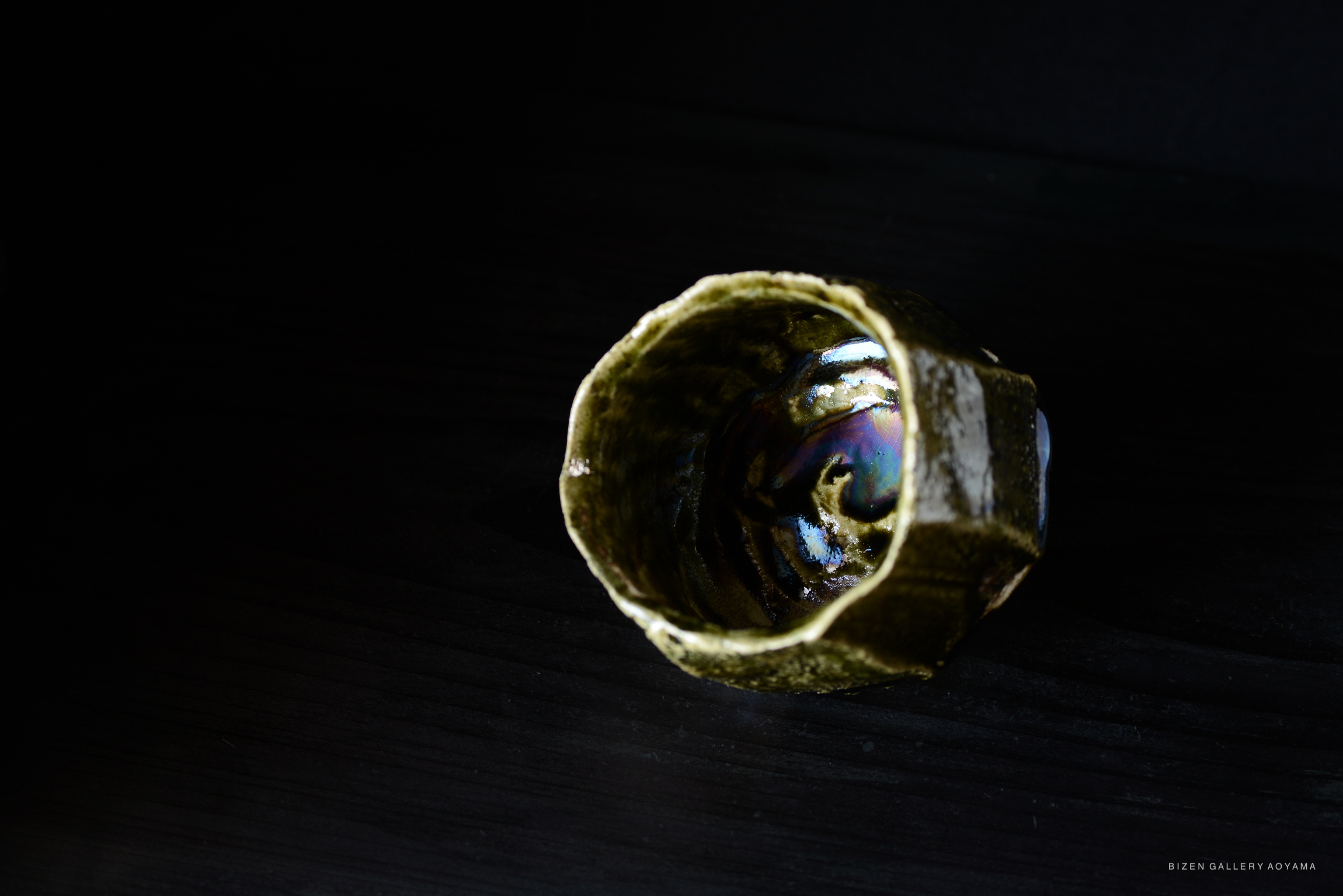 A top-down view of an intricately crafted ceramic cup with a textured surface and iridescent glimmering interior, set against a dark background.