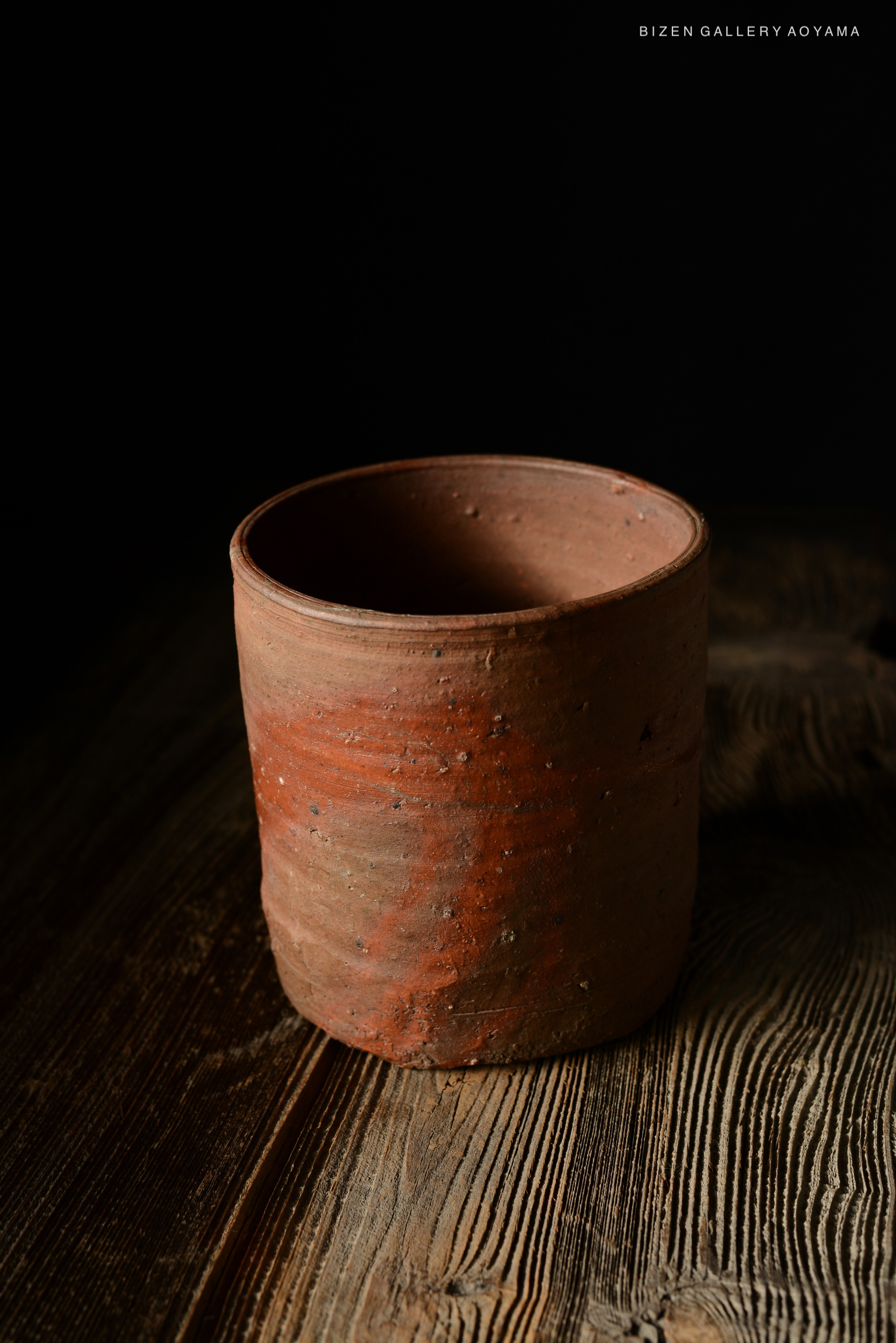 A close-up of a cylindrical, unglazed Bizen ware pot displayed on a wooden surface with a dark background.