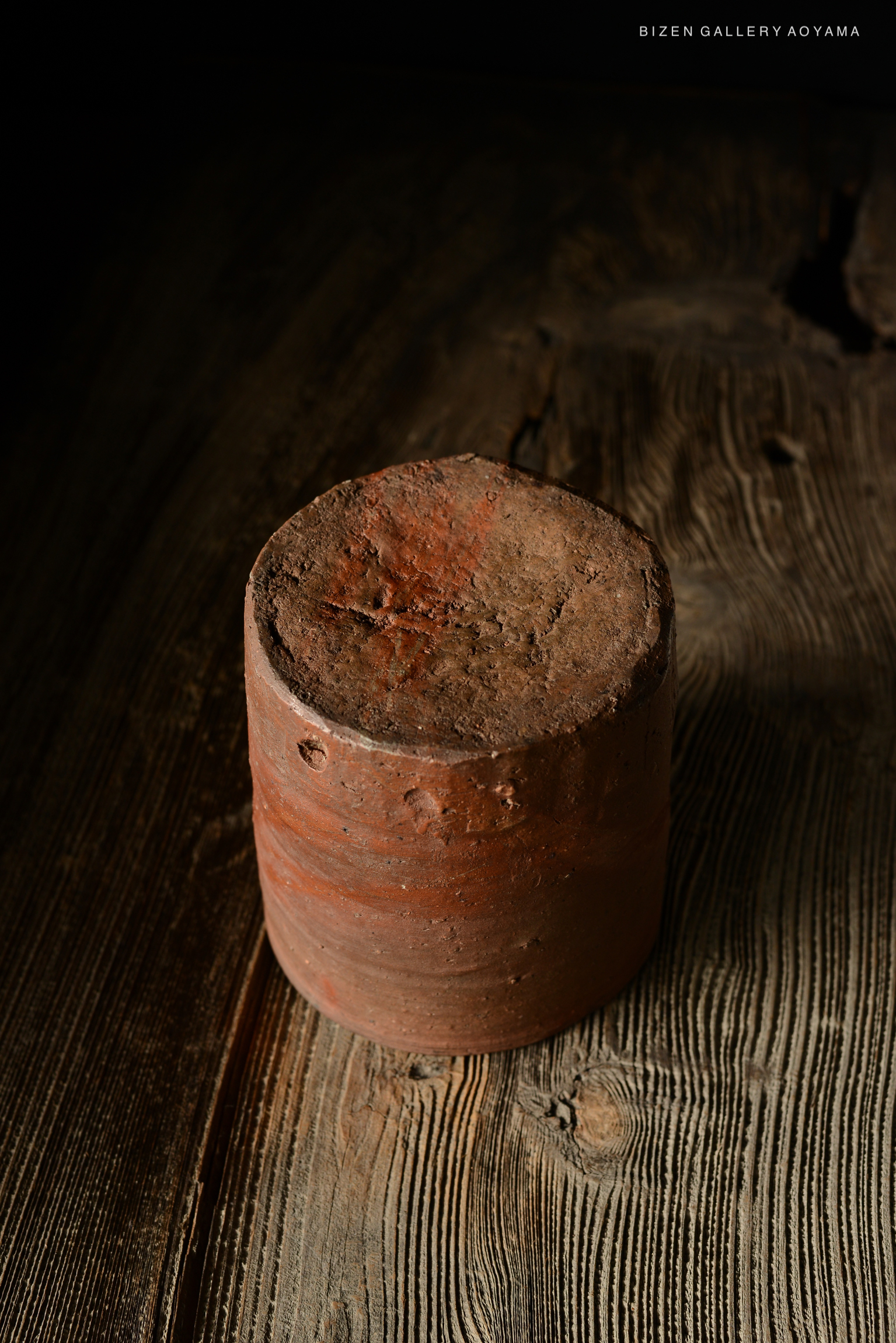 A close-up view of a cylindrical, rustic pottery piece with a rough, textured surface, sitting on a wooden surface.