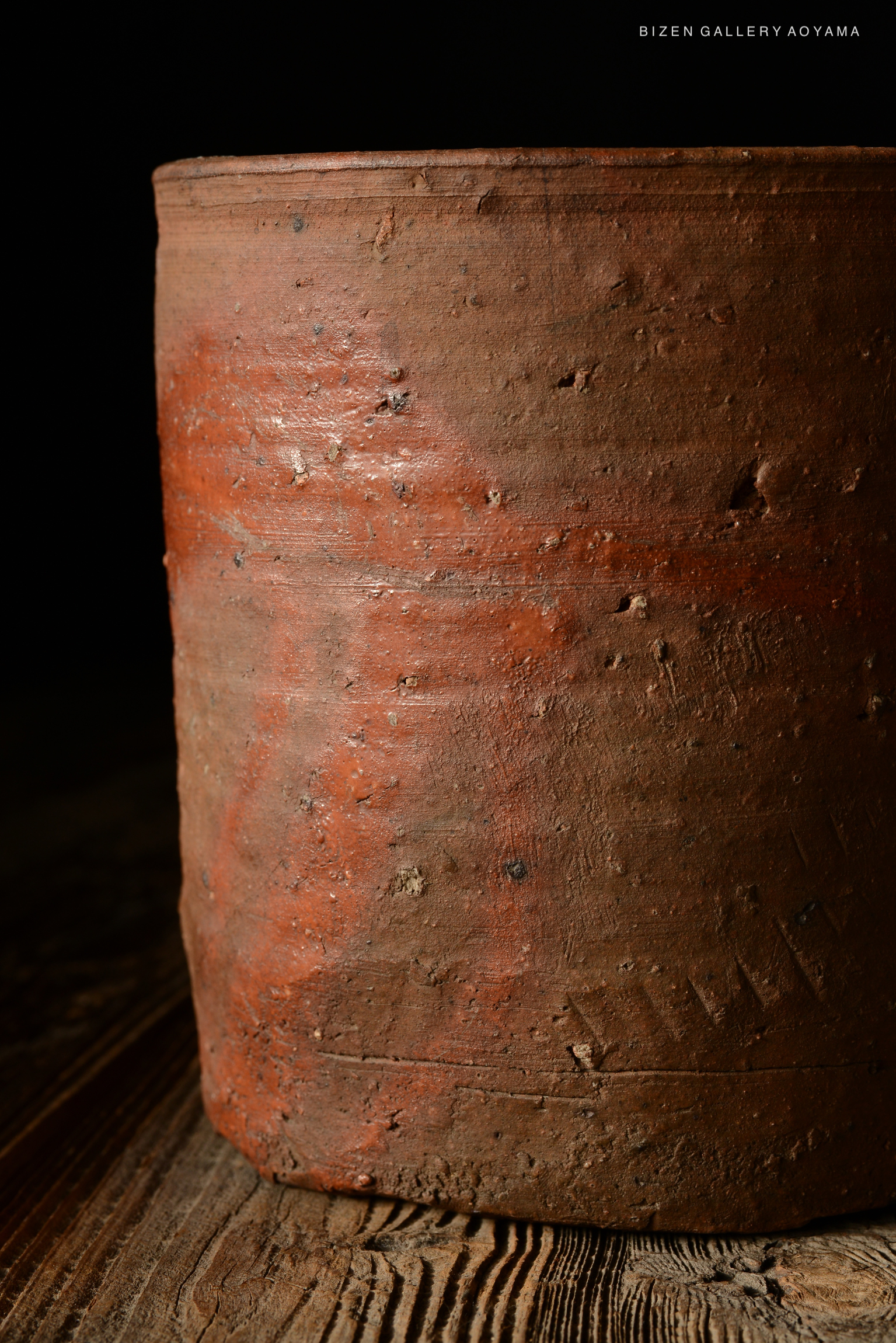 Close-up of a rustic, unglazed Bizen pottery piece with a textured, reddish surface, set against a dark background.