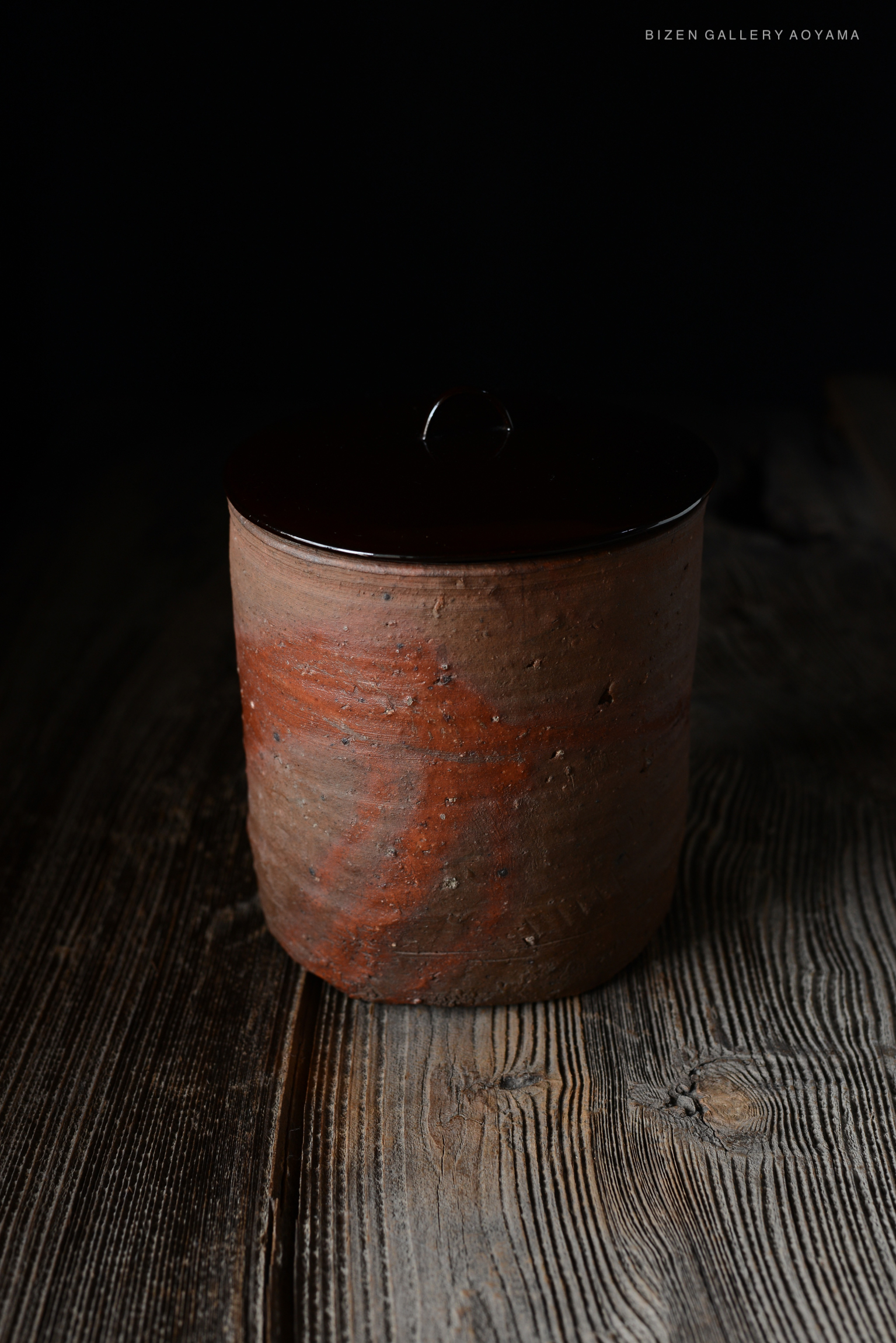 A round Bizen pottery water container with a glossy dark lid, placed on a rustic wooden surface. The pot features a subtle reddish hue and a textured finish, indicative of craftsmanship from the Momoyama to early Edo period.