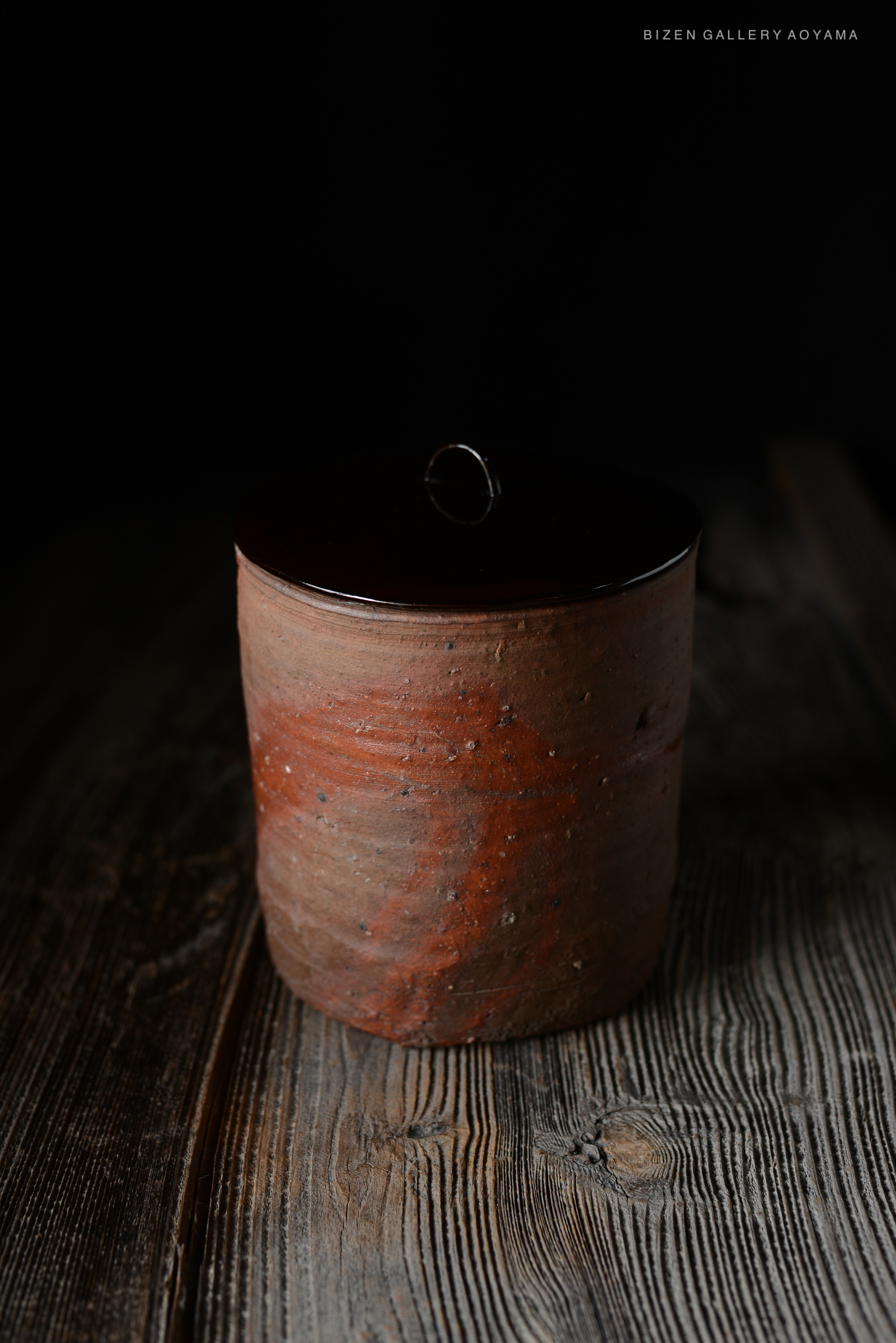 A rustic Bizen pottery mizusashi with a simple cylindrical shape and a dark, glossy lid, displayed on a wooden surface.