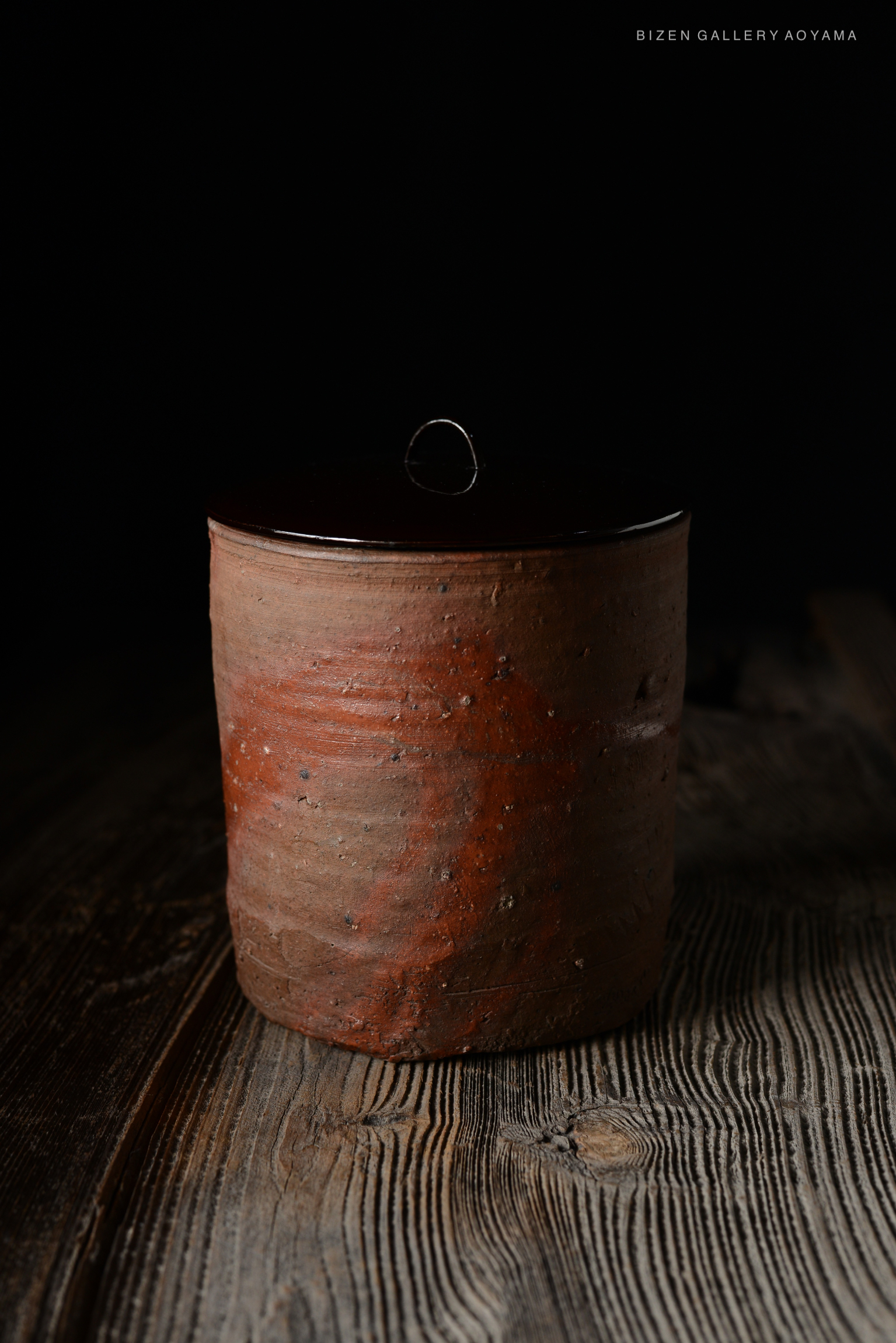 A traditional Japanese tea container made of unglazed clay, featuring a rounded body with a deep reddish-brown tint and a glossy black lid, placed on a rustic wooden surface.