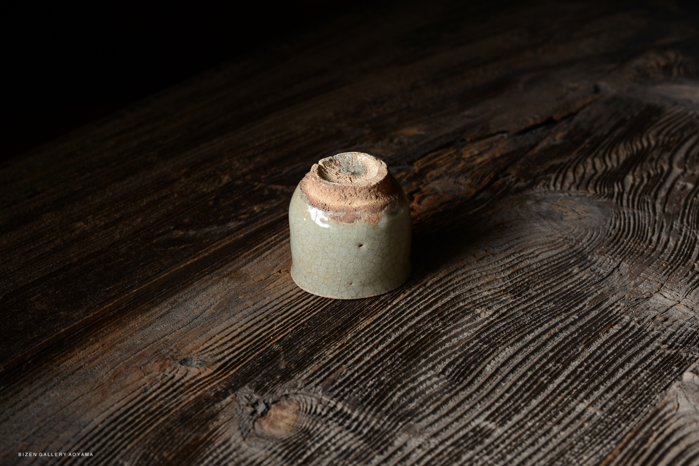 A small, cracked green ceramic vessel placed on a textured wooden surface.