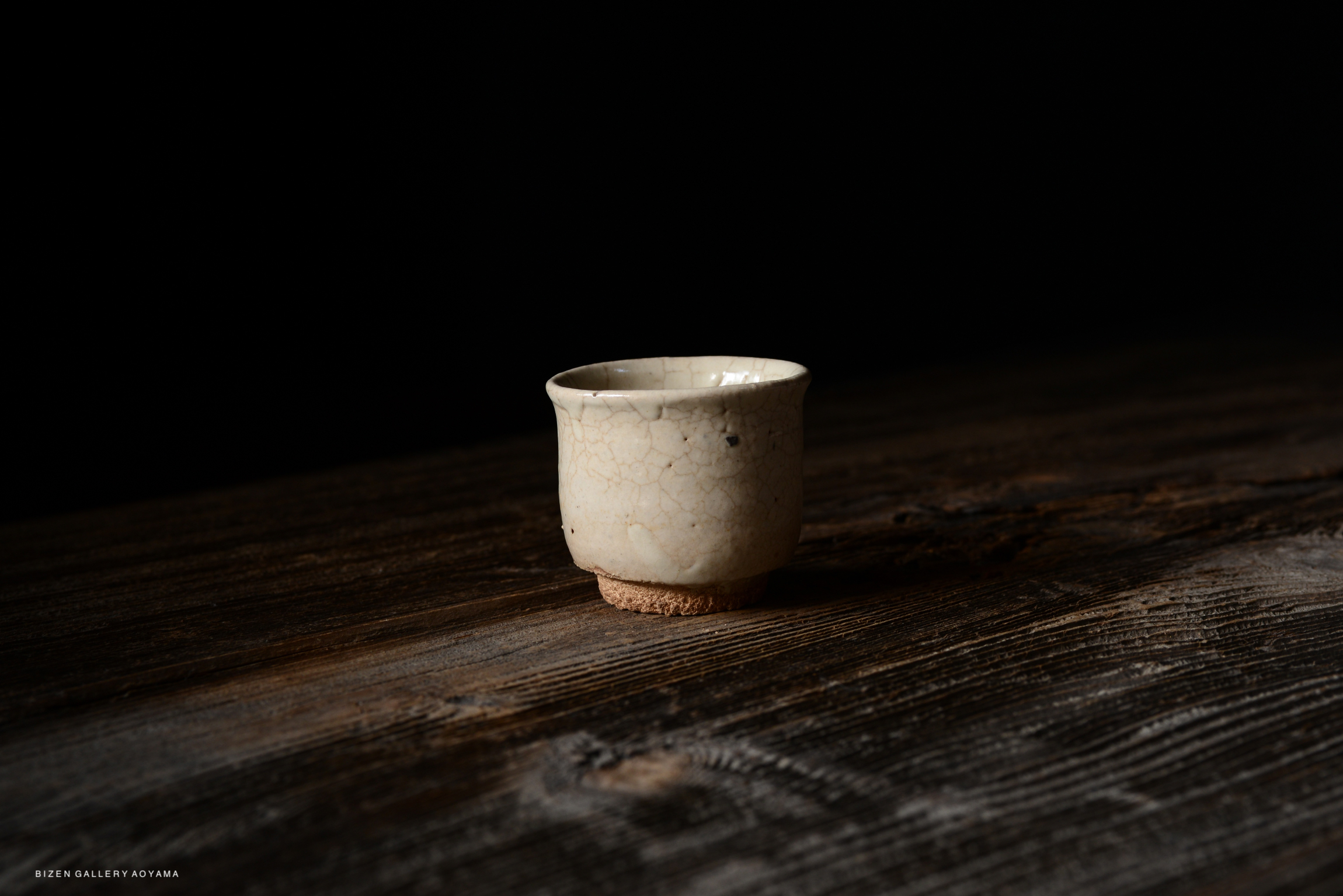 A small, cracked ceramic cup resting on a wooden surface, with a dark background.
