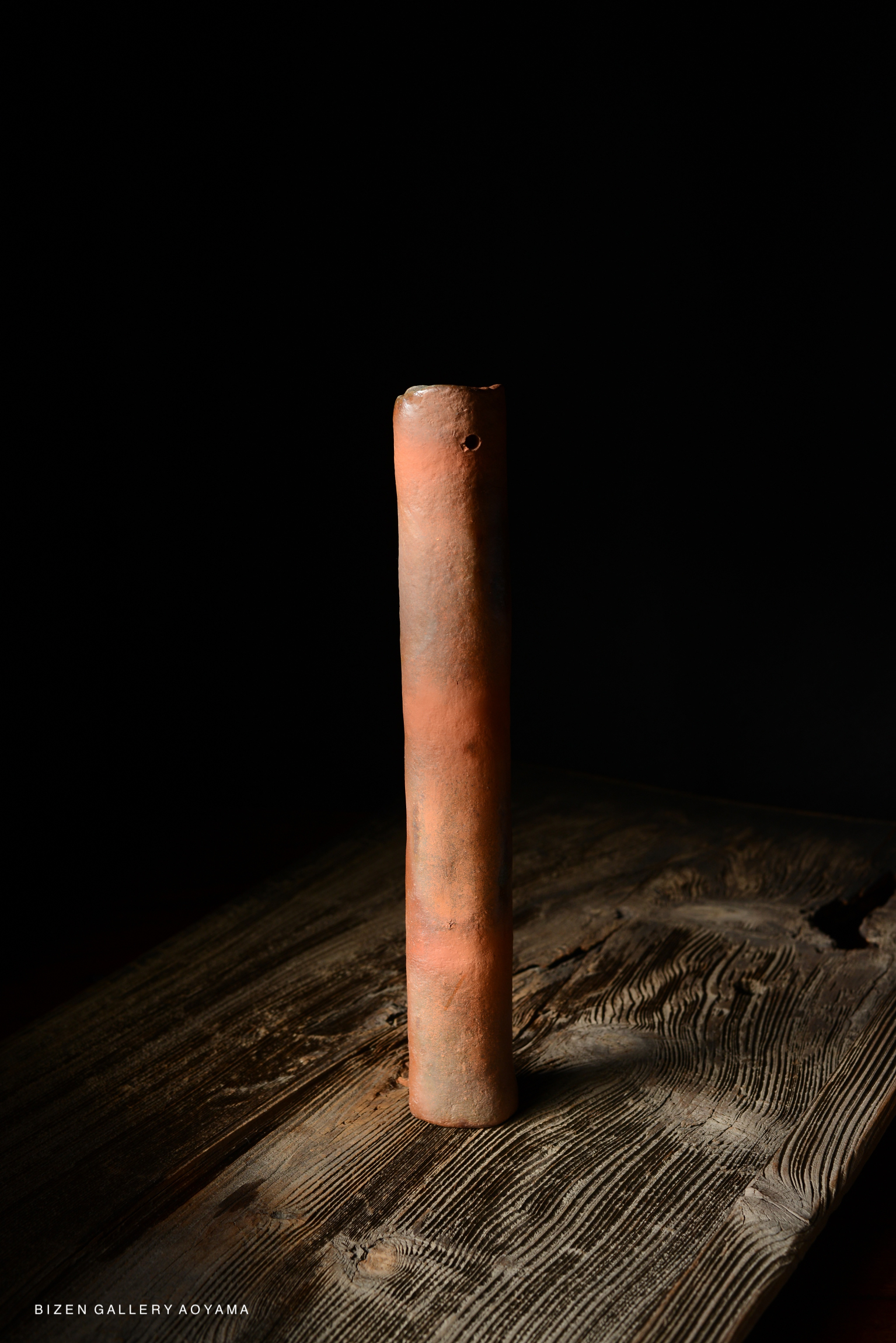 A tall, cylindrical Bizen pottery vase with a muted earthy color, displayed against a dark background on a textured wooden surface.