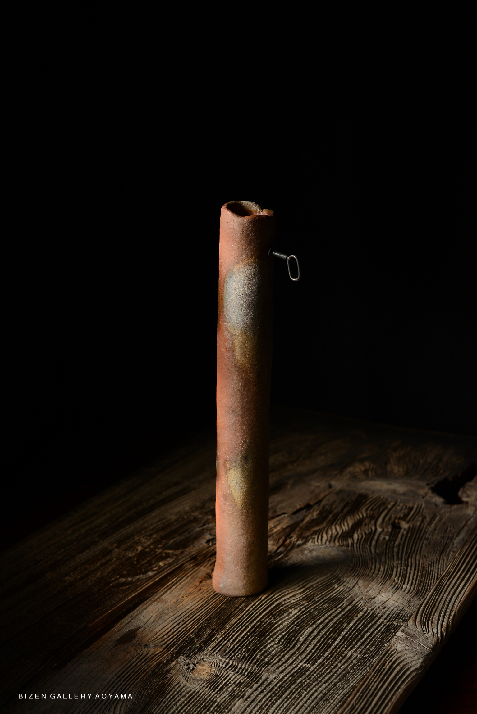 A tall, cylindrical ceramic vase with a textured surface and a small metal handle, set against a dark background.