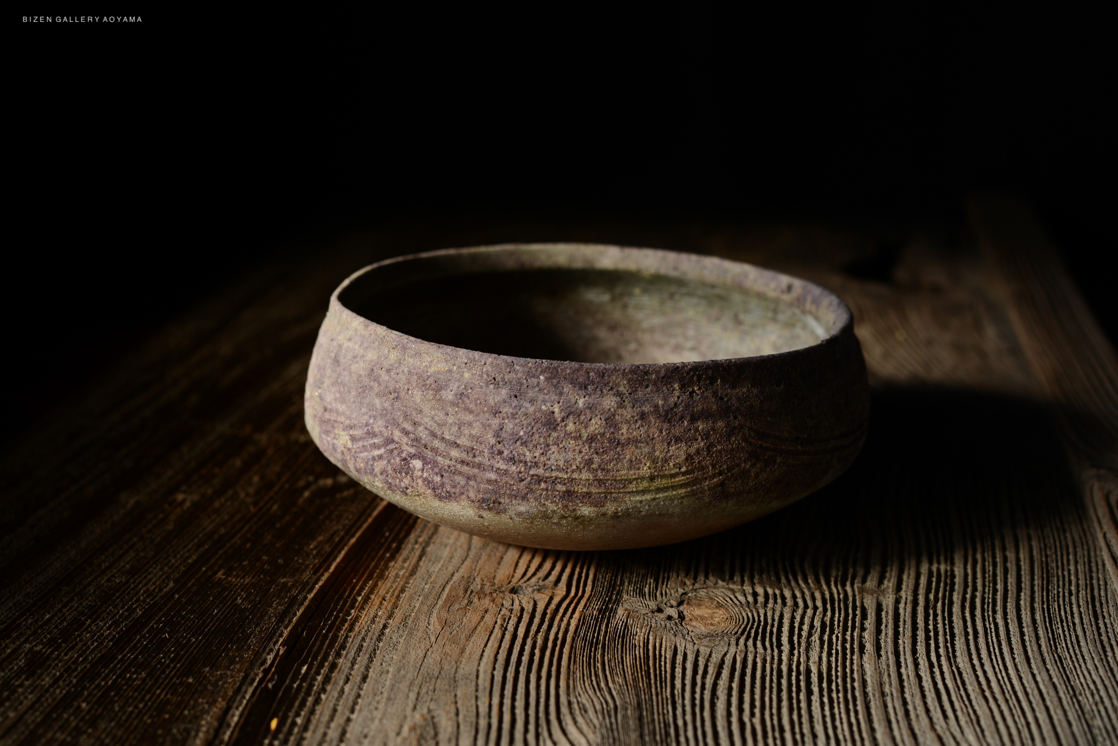 A rustic ceramic bowl with a textured surface, set on a wooden table, partially illuminated against a dark background.