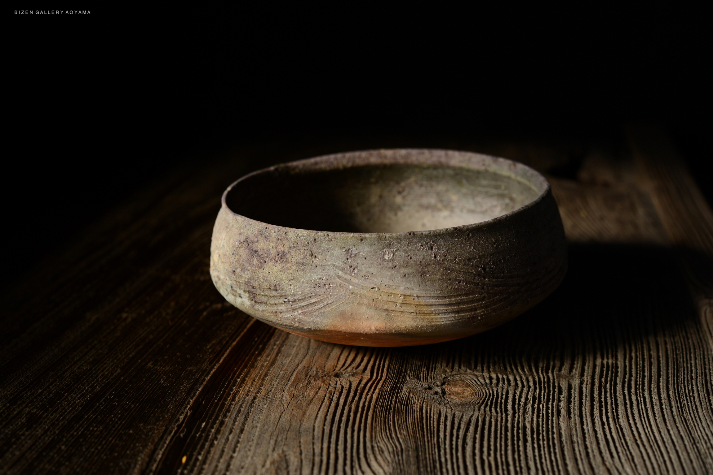 A weathered ceramic bowl placed on a wooden surface, illuminated by soft lighting, showcasing its textures and colors against a dark background.