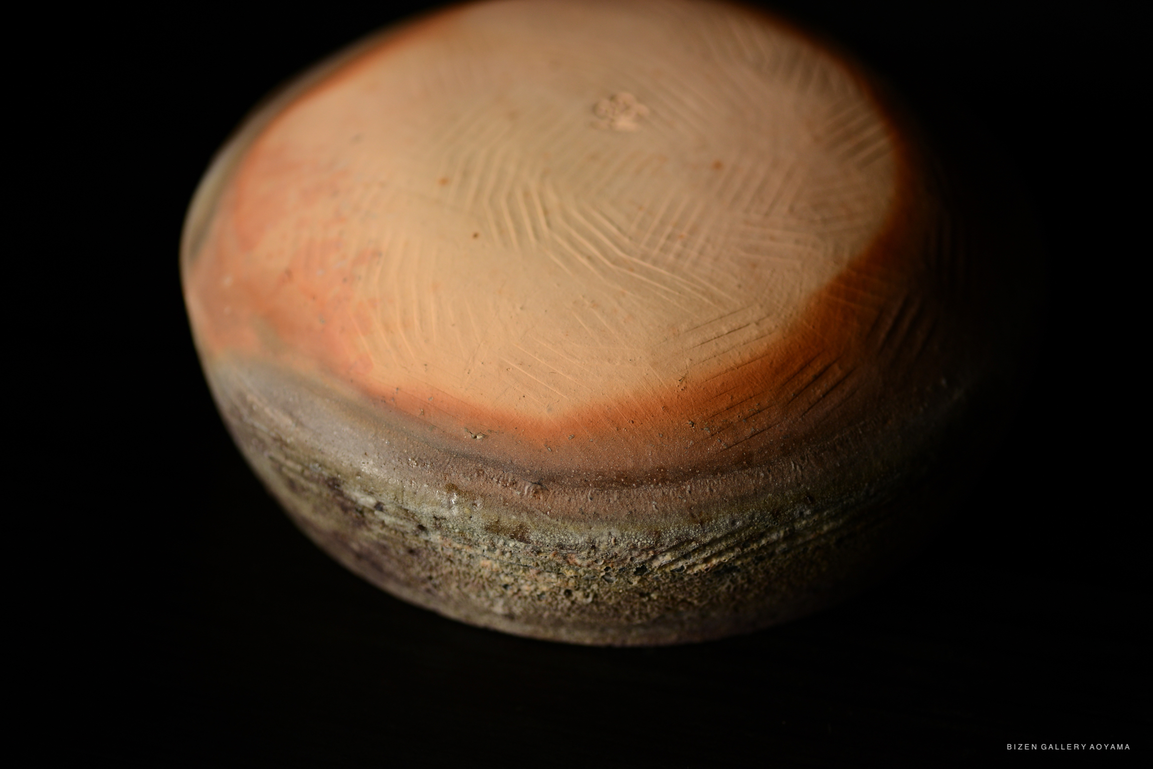 A close-up of a round, earthy ceramic bowl with a smooth, patterned surface and a rustic edge, set against a dark background.