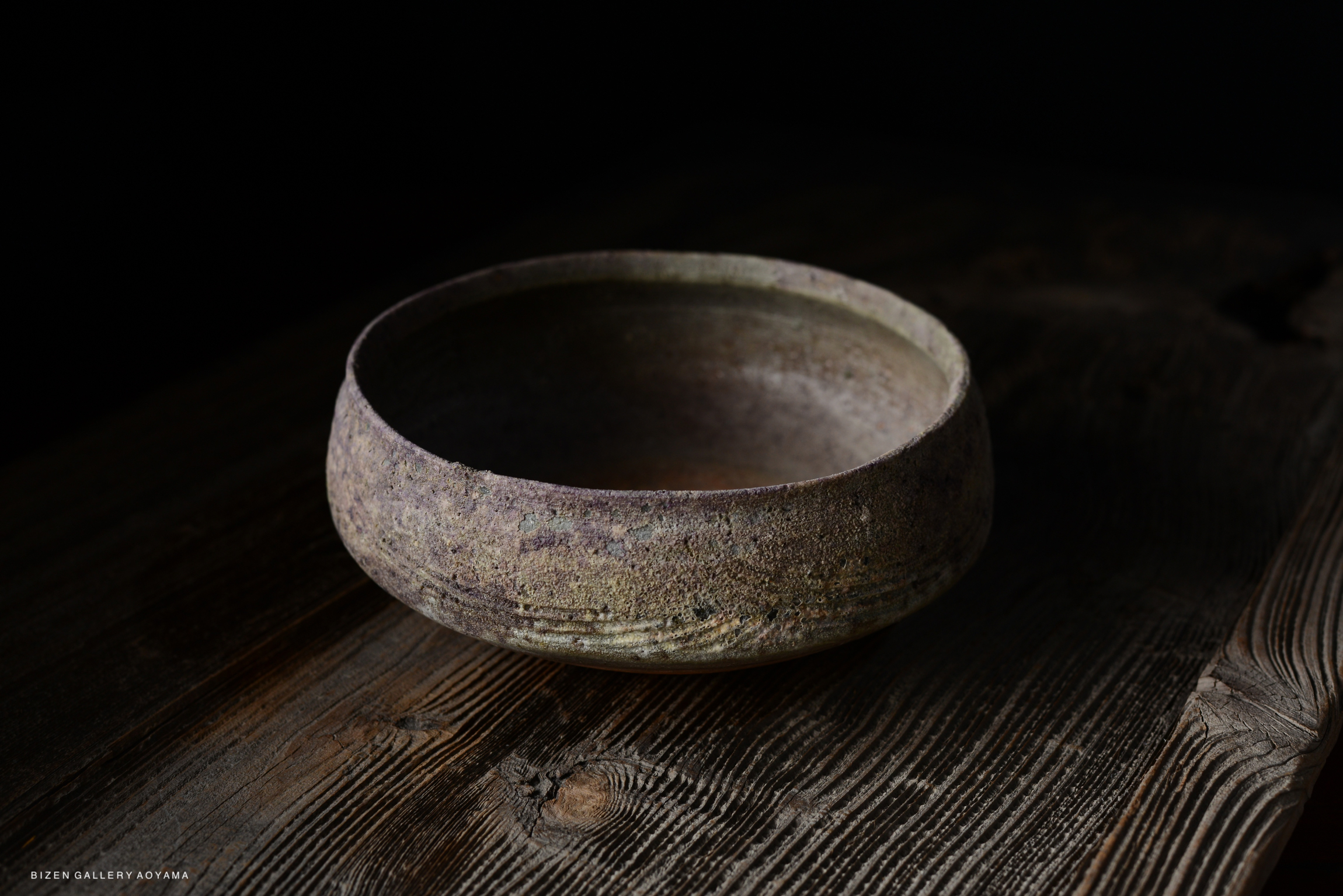 A rustic Bizen Haiki tea ceremony vessel with a textured surface in shades of purple and scarlet, placed on a wooden table.