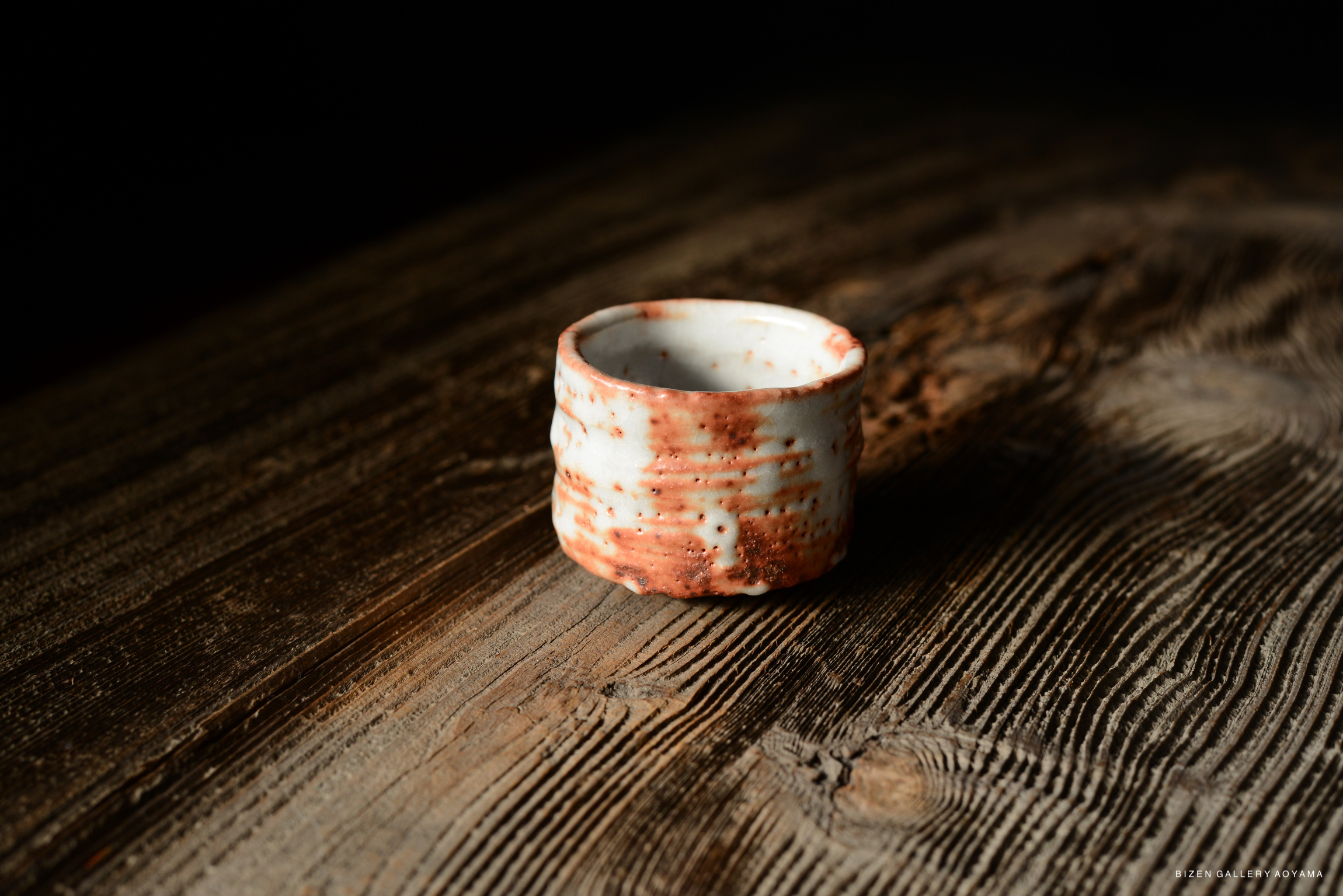 A small ceramic cup with a textured surface and reddish-brown accents, placed on a rustic wooden table.