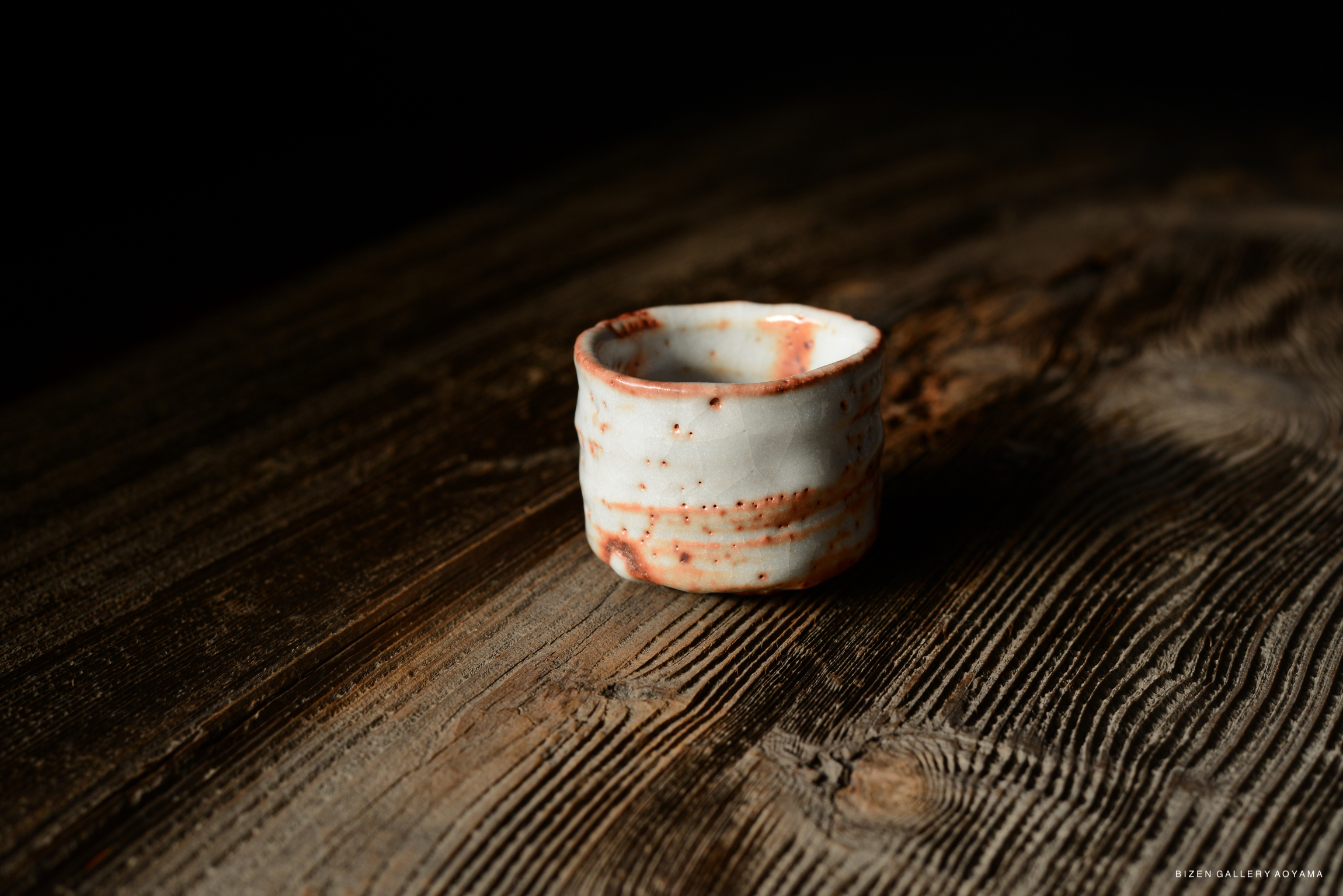 A small, rustic ceramic cup with a white glaze and reddish-brown accents, sitting on a textured wooden surface.