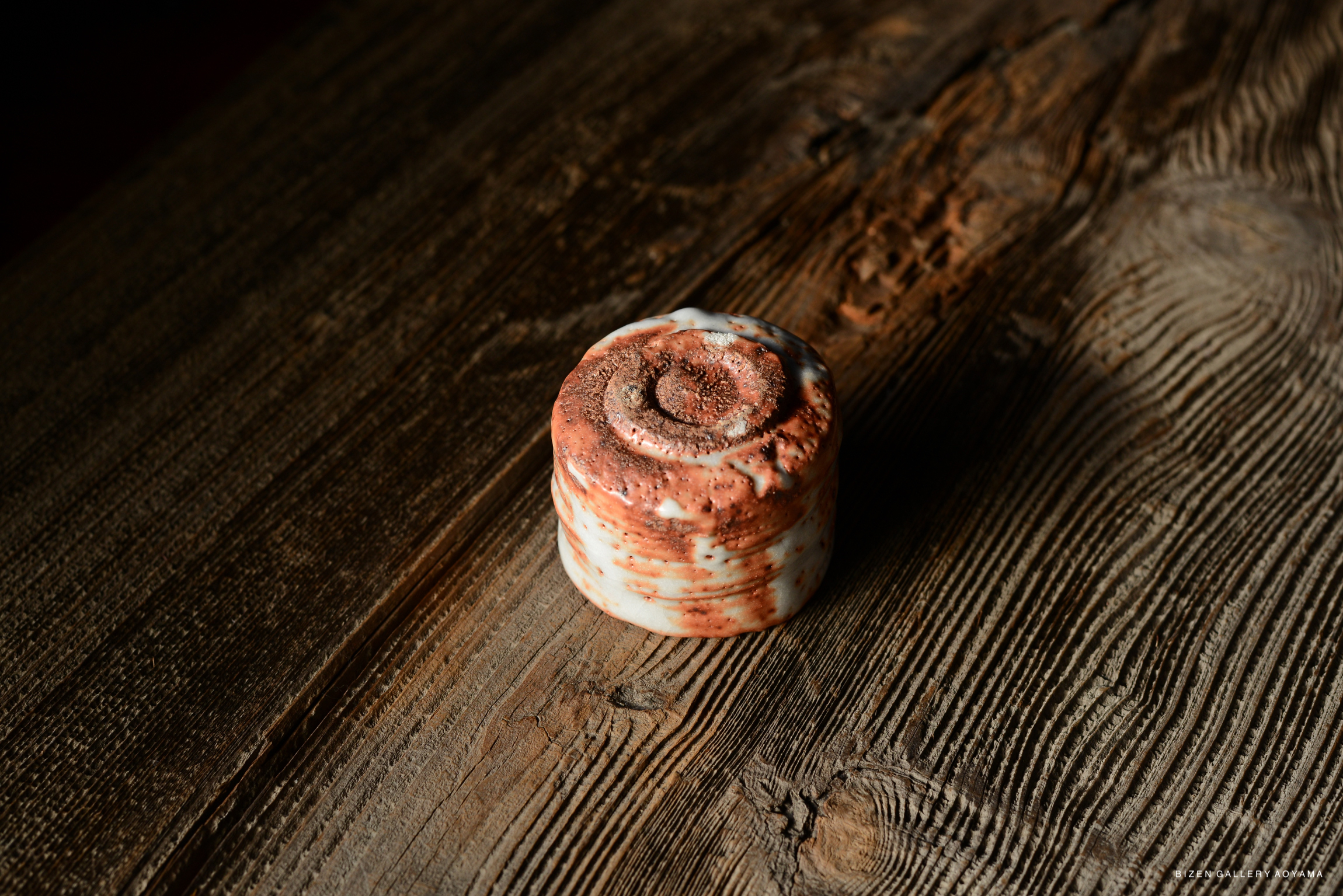 A close-up view of a round ceramic container with a speckled red and white glaze, resting on a textured wooden surface.