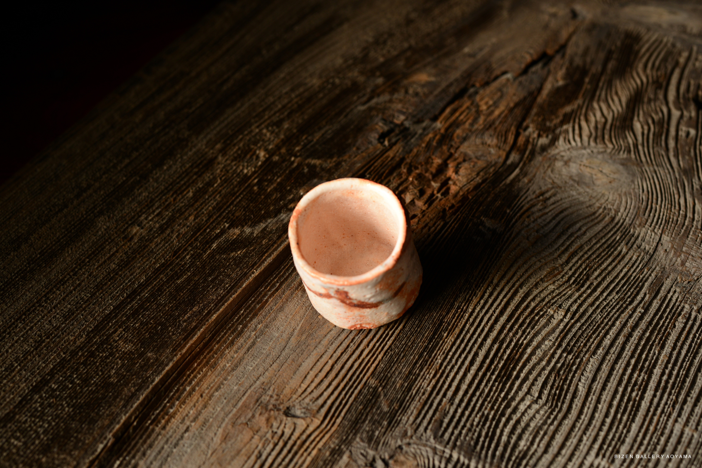 A close-up view of a handcrafted Shino Guinomi sake cup resting on a textured wooden surface.