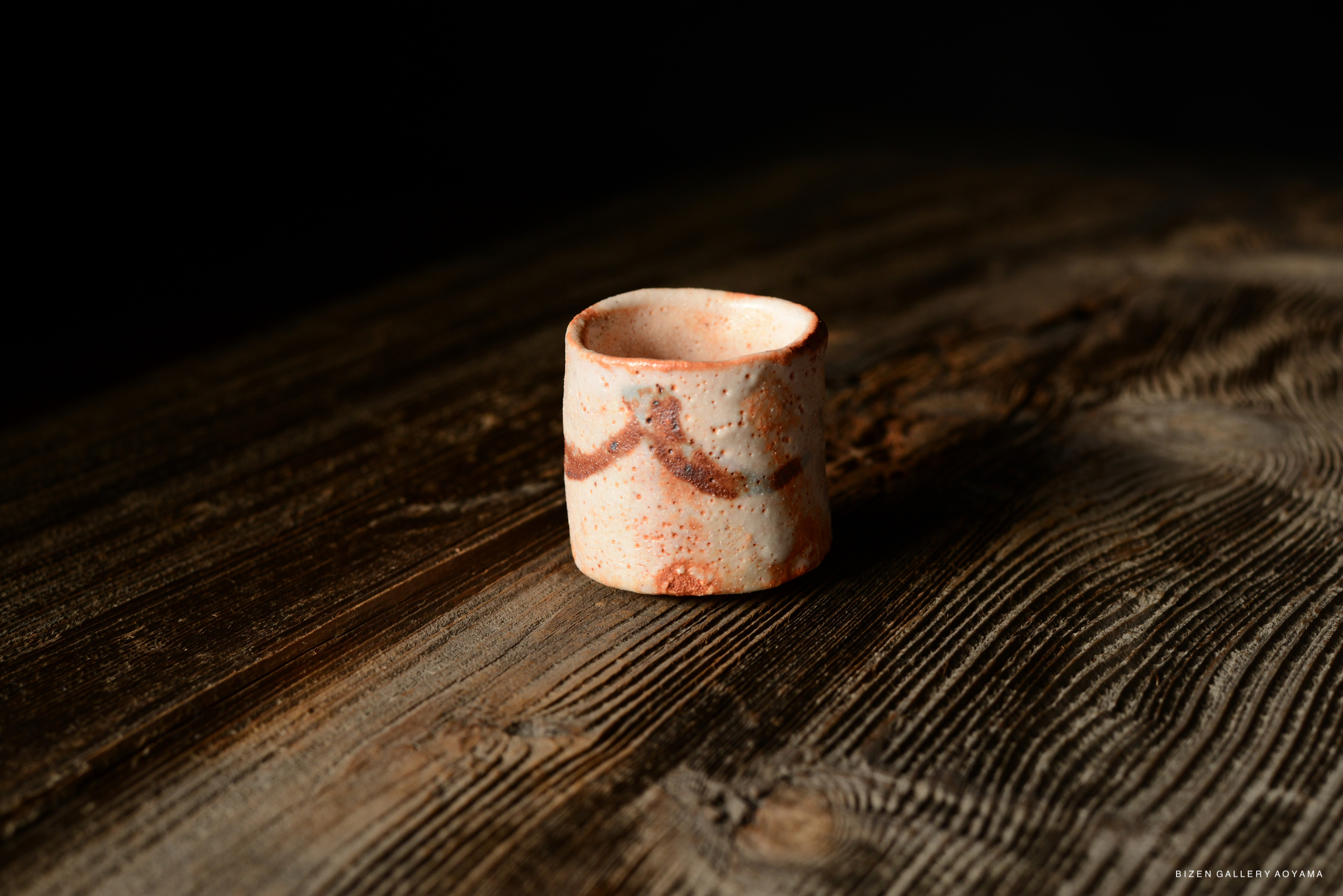 A Shino Guinomi, a traditional Japanese drinking vessel, resting on a textured wooden surface. The cup features a decorative pattern and a soft color palette.