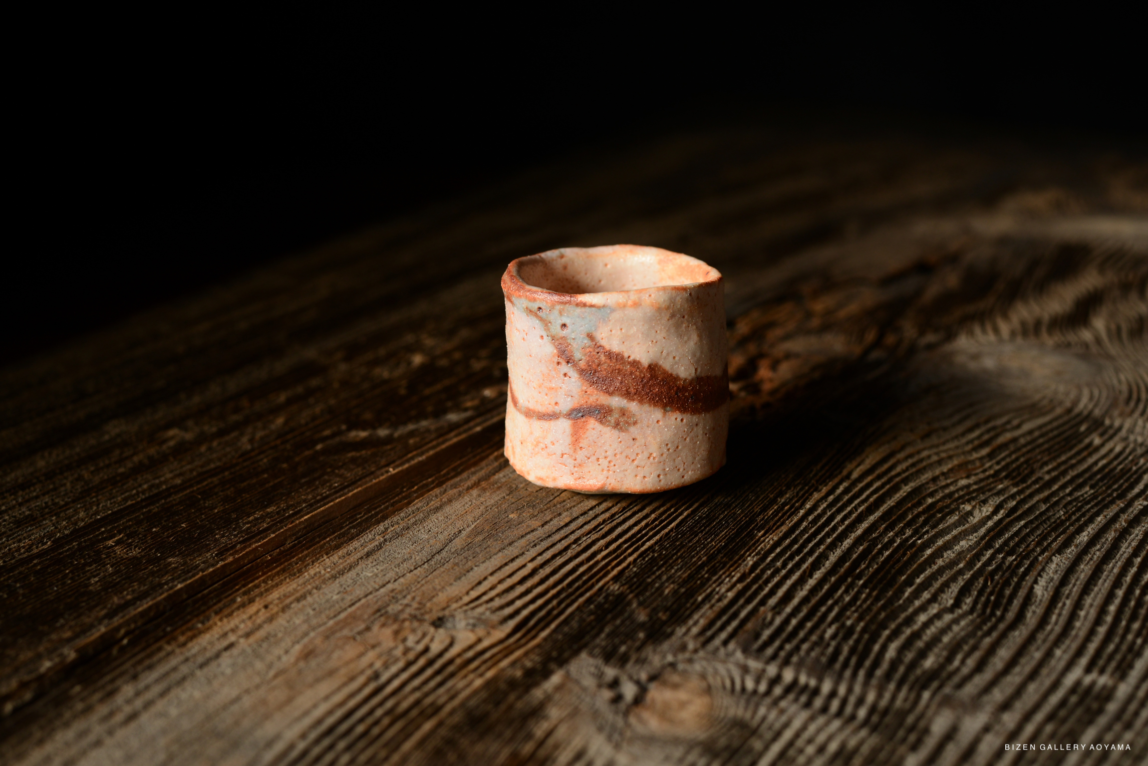 A close-up image of a Shino Guinomi sake cup with a textured surface, featuring earthy tones and patterns, placed on a rustic wooden table.